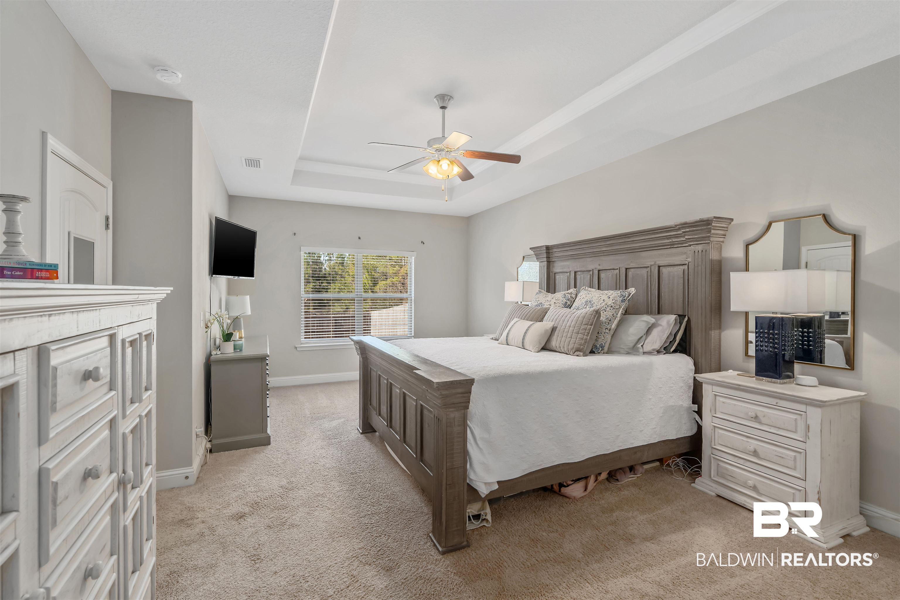 Primary bedroom with a large dark wood bed, tray ceiling, ceiling fan, and window with blinds.