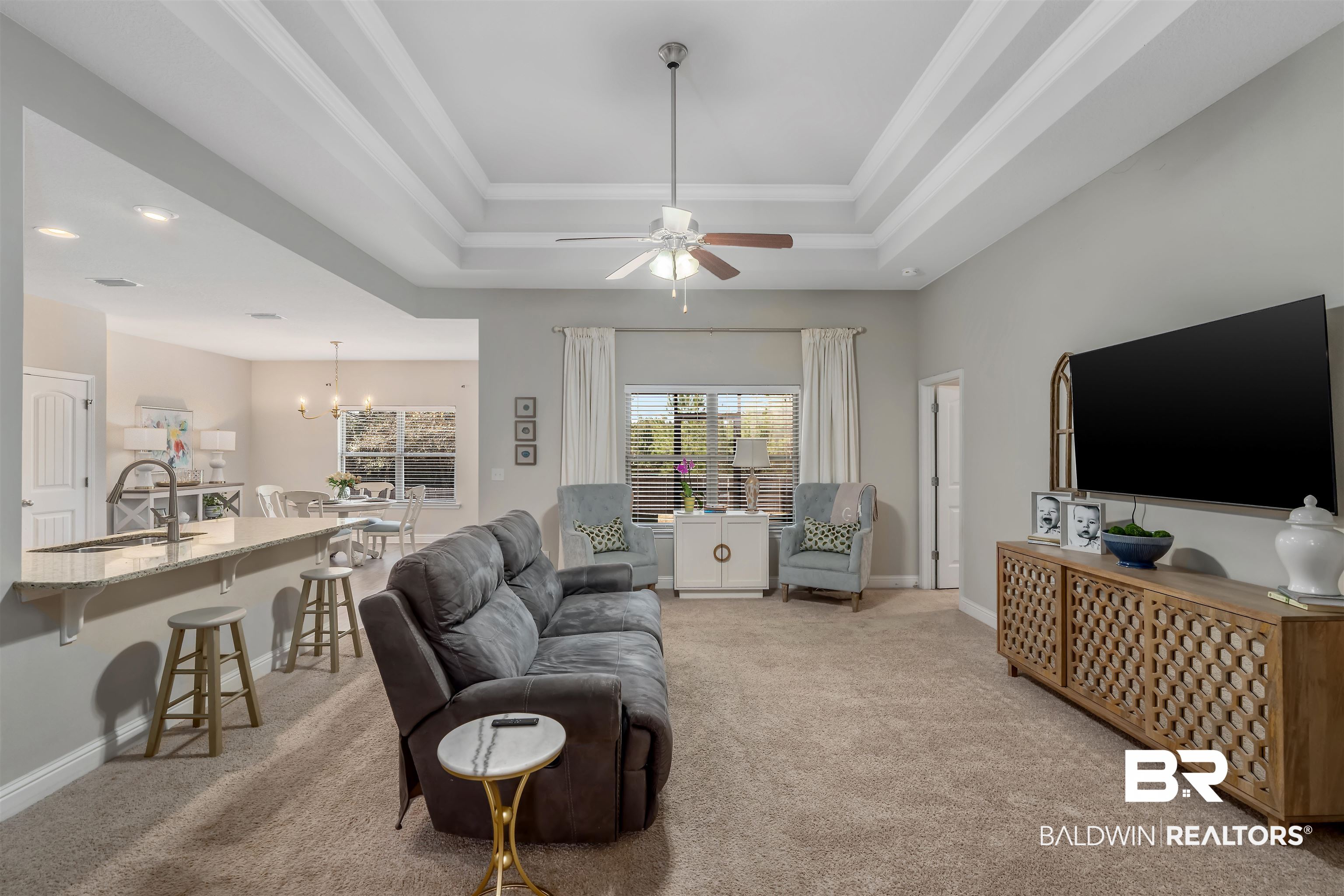 Open-plan living room with gray sofa, armchairs, tray ceiling, and adjoining dining area and kitchen.