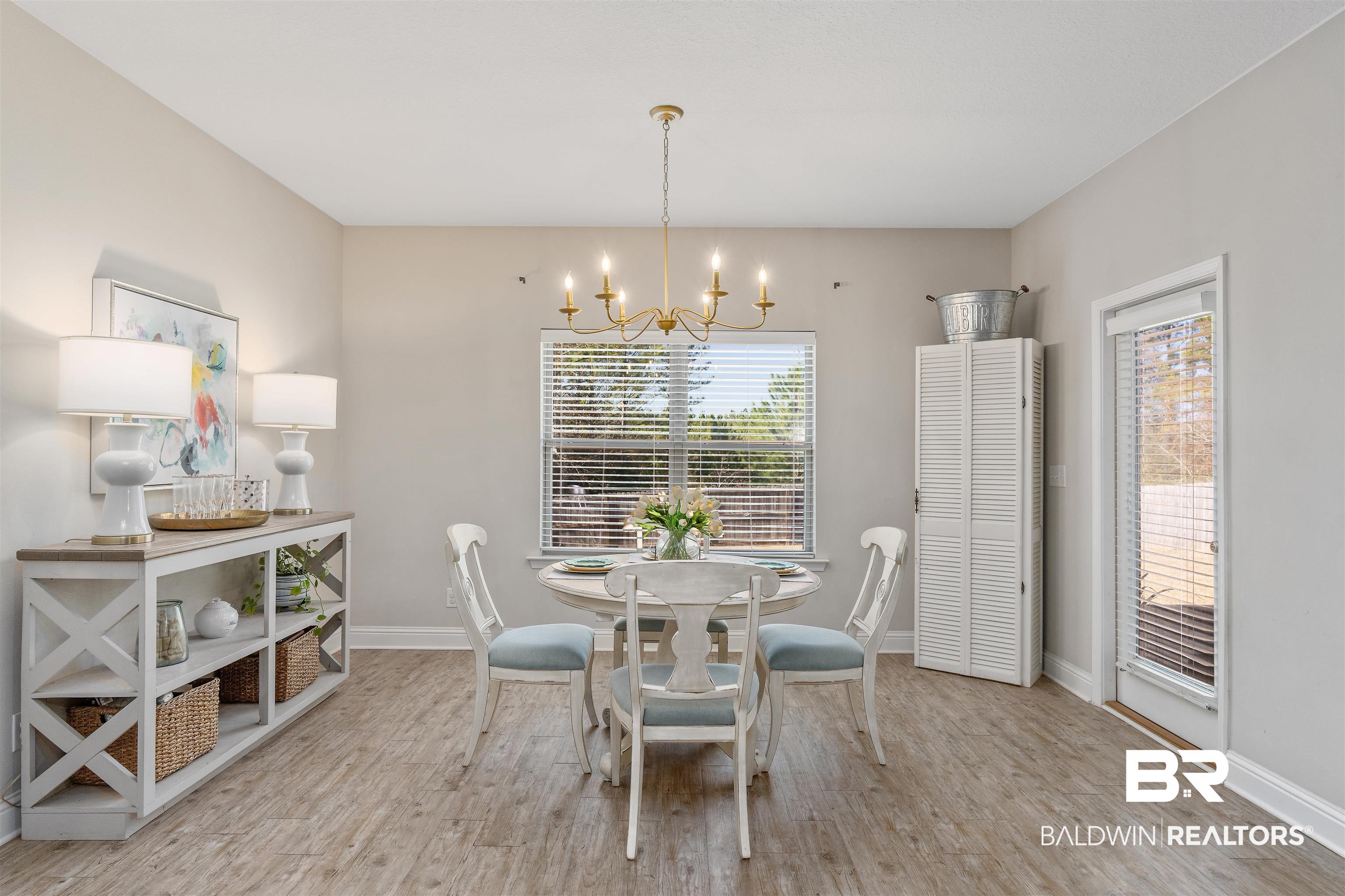 Furnished dining room with a round table, brass chandelier, and large windows with blinds.