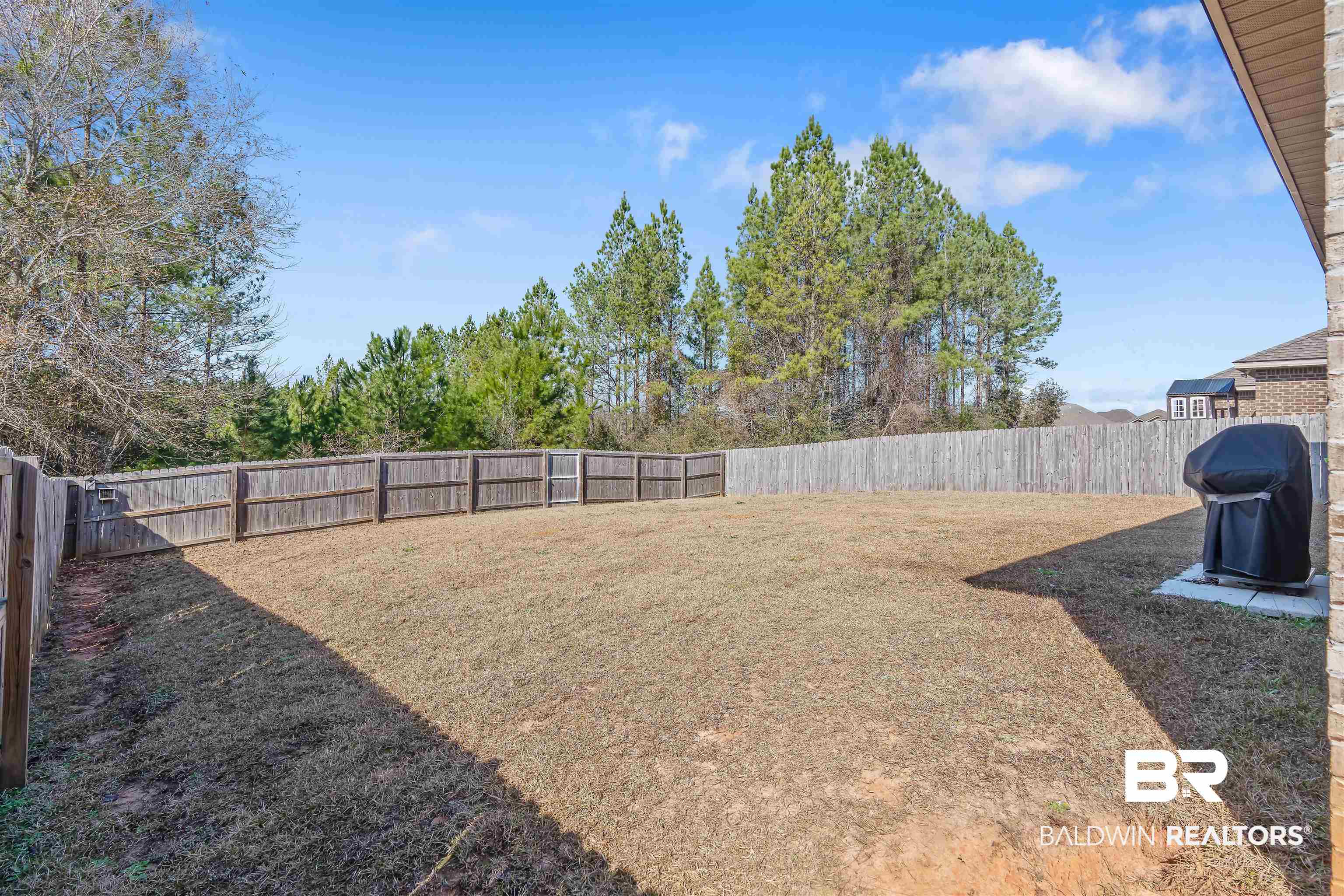 Fenced backyard with a broad lawn, mature trees, and a covered grill beside the house.