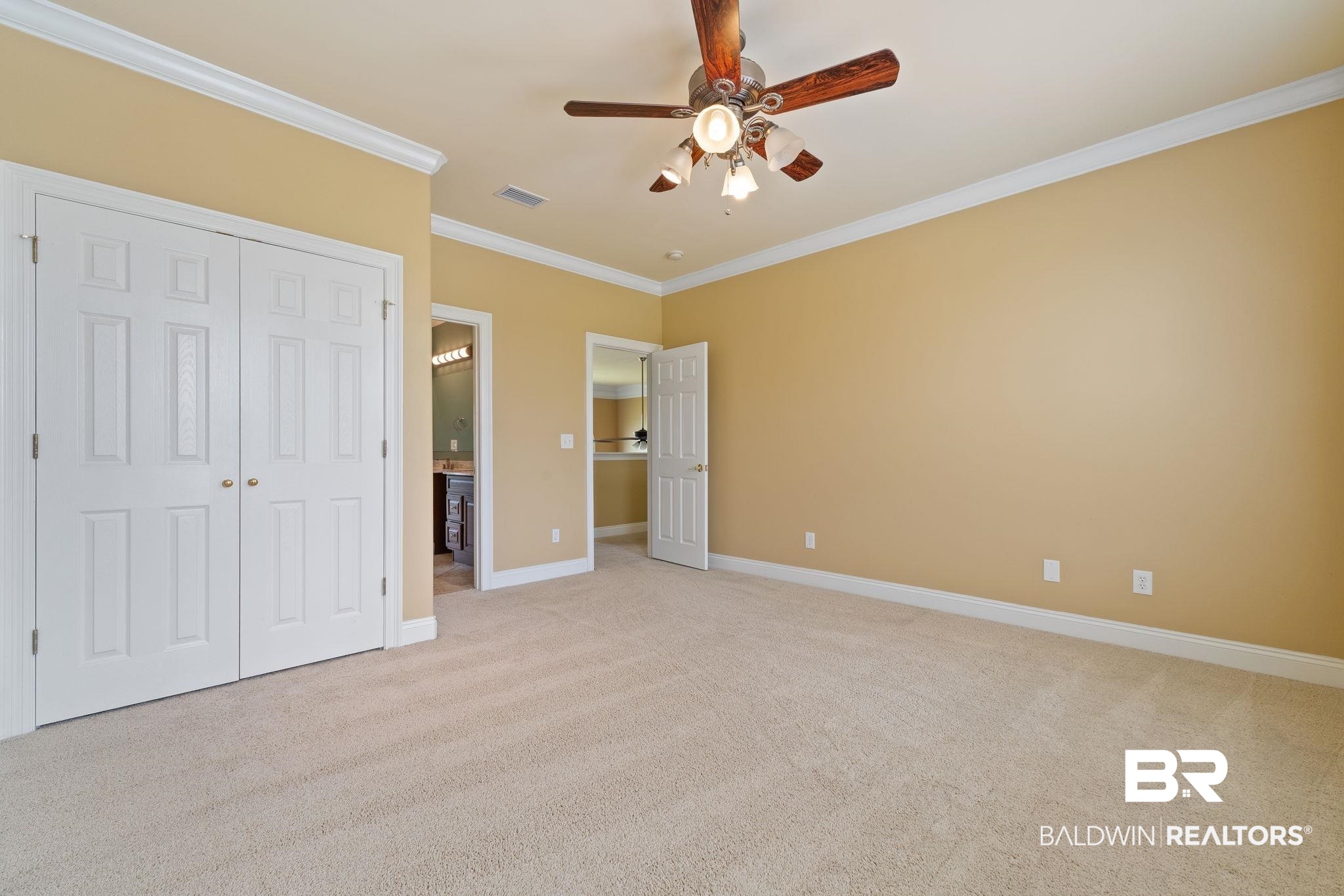 Empty beige-carpeted bedroom with double closets, ceiling fan, and open doors to adjoining rooms.