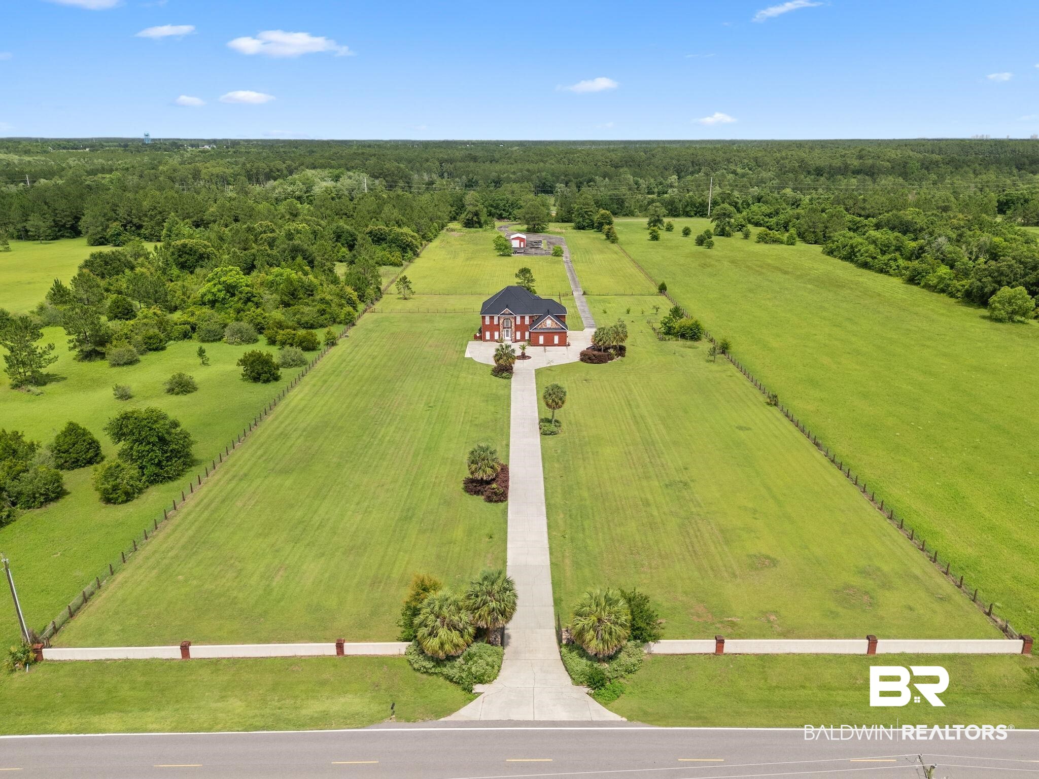 Aerial view of a red-brick house with a long driveway on a large fenced lawn.