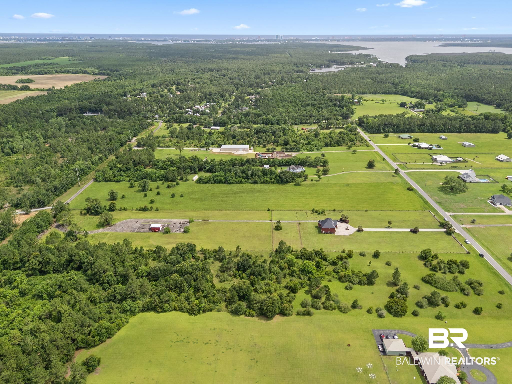 Aerial view of rural acreage with open fields, scattered homes, woods, and distant water.