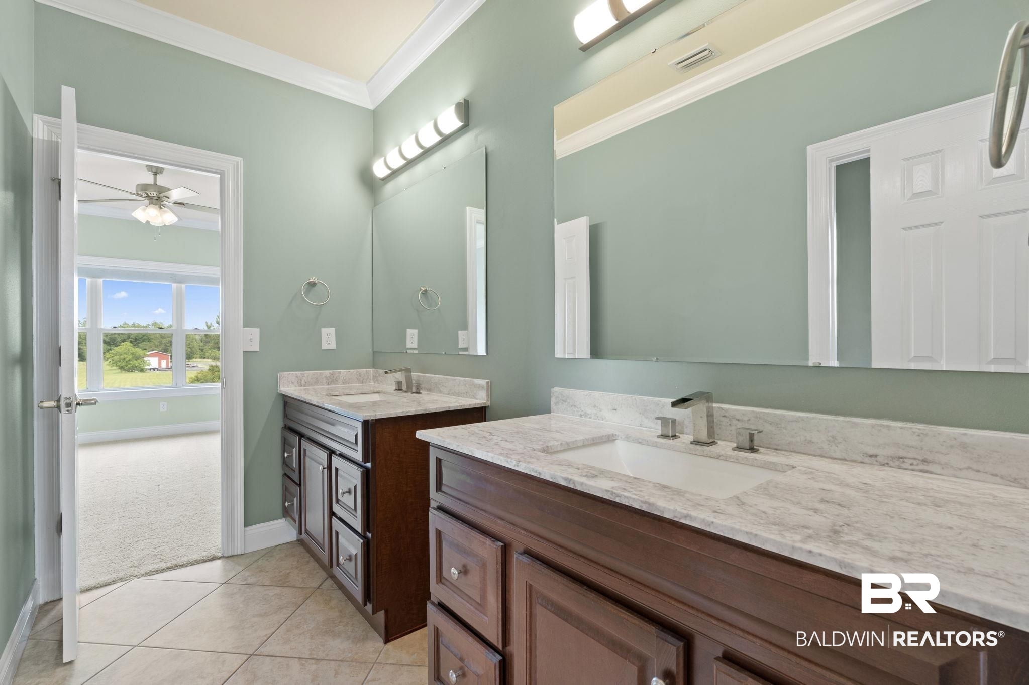 Bathroom with double wood vanities, stone countertops, and large mirrors beside an open doorway.