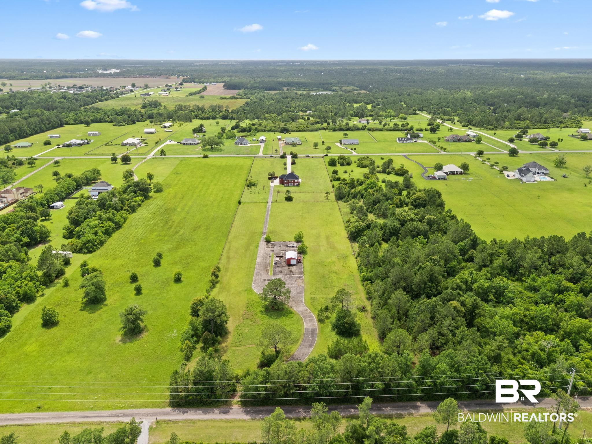 Aerial view of a rural property with a long driveway, central house, outbuilding, open lawn, and surrounding woods.