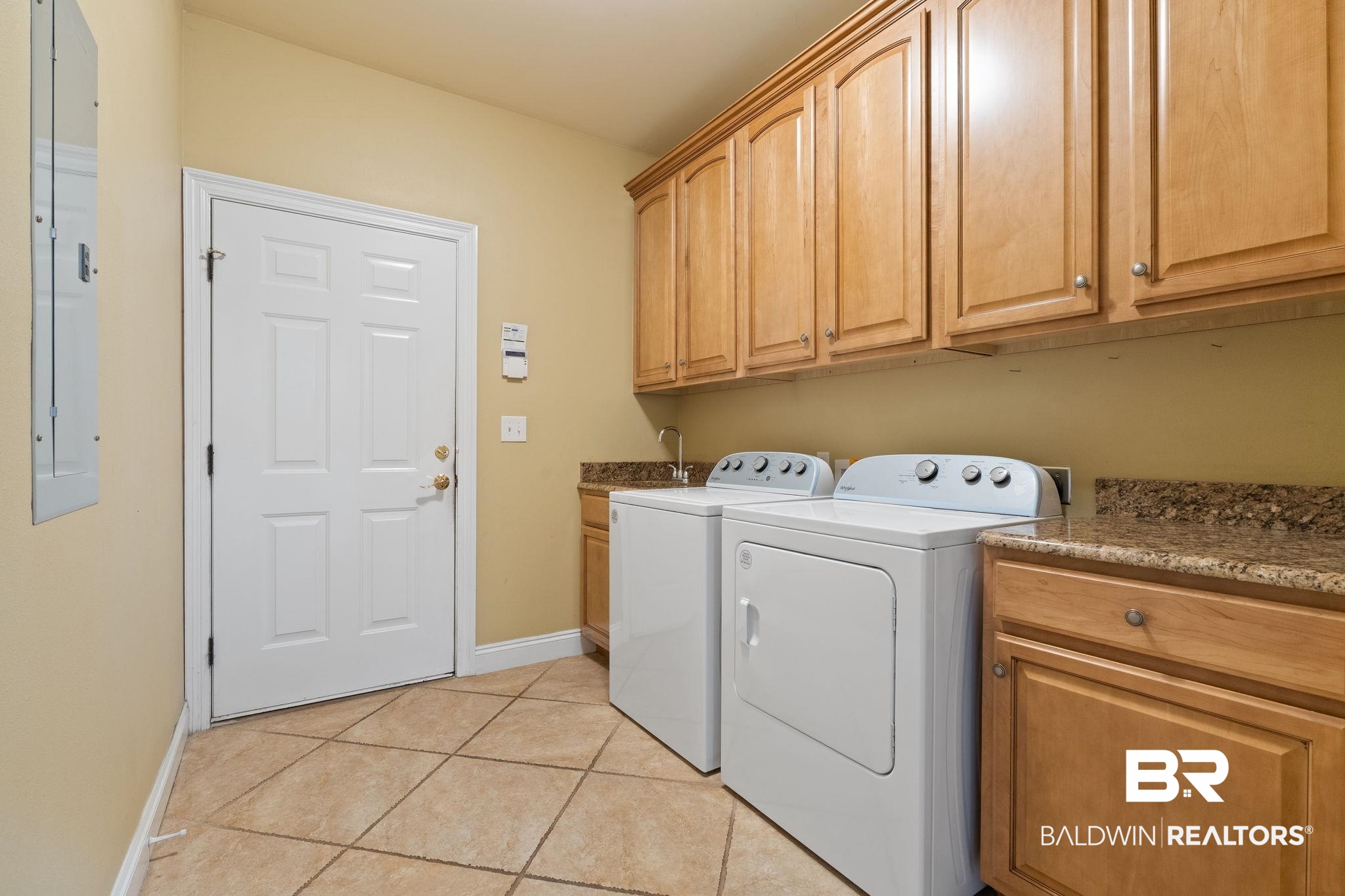 Laundry room with white washer and dryer, wood cabinetry, speckled countertops, and tan tile flooring.