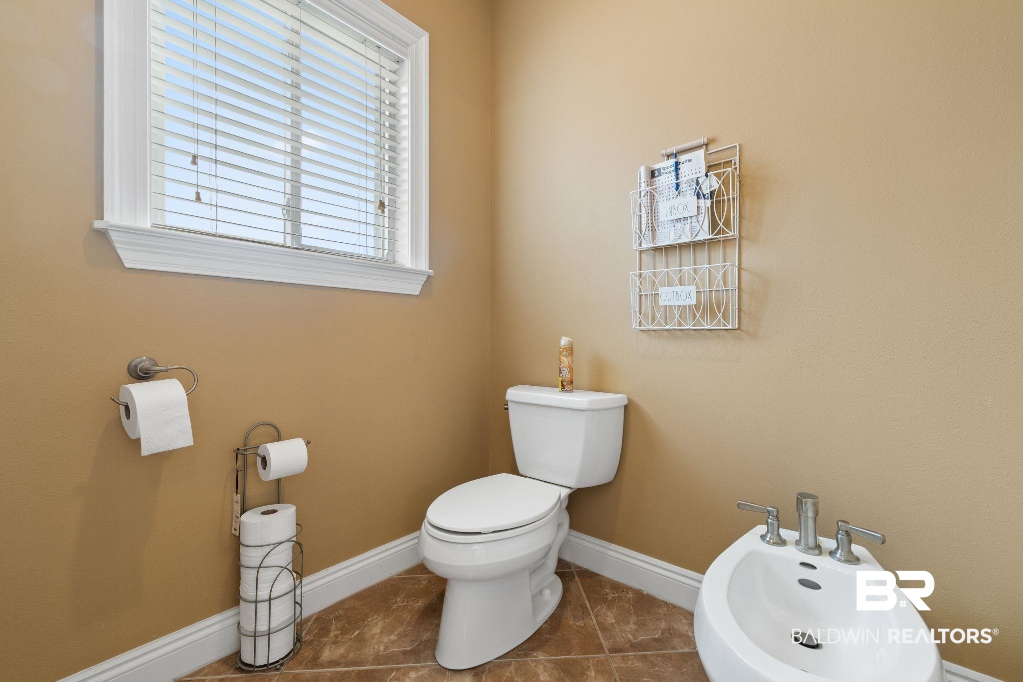 Bathroom with a white toilet and bidet, tan walls, brown tile floor, and a window with blinds.