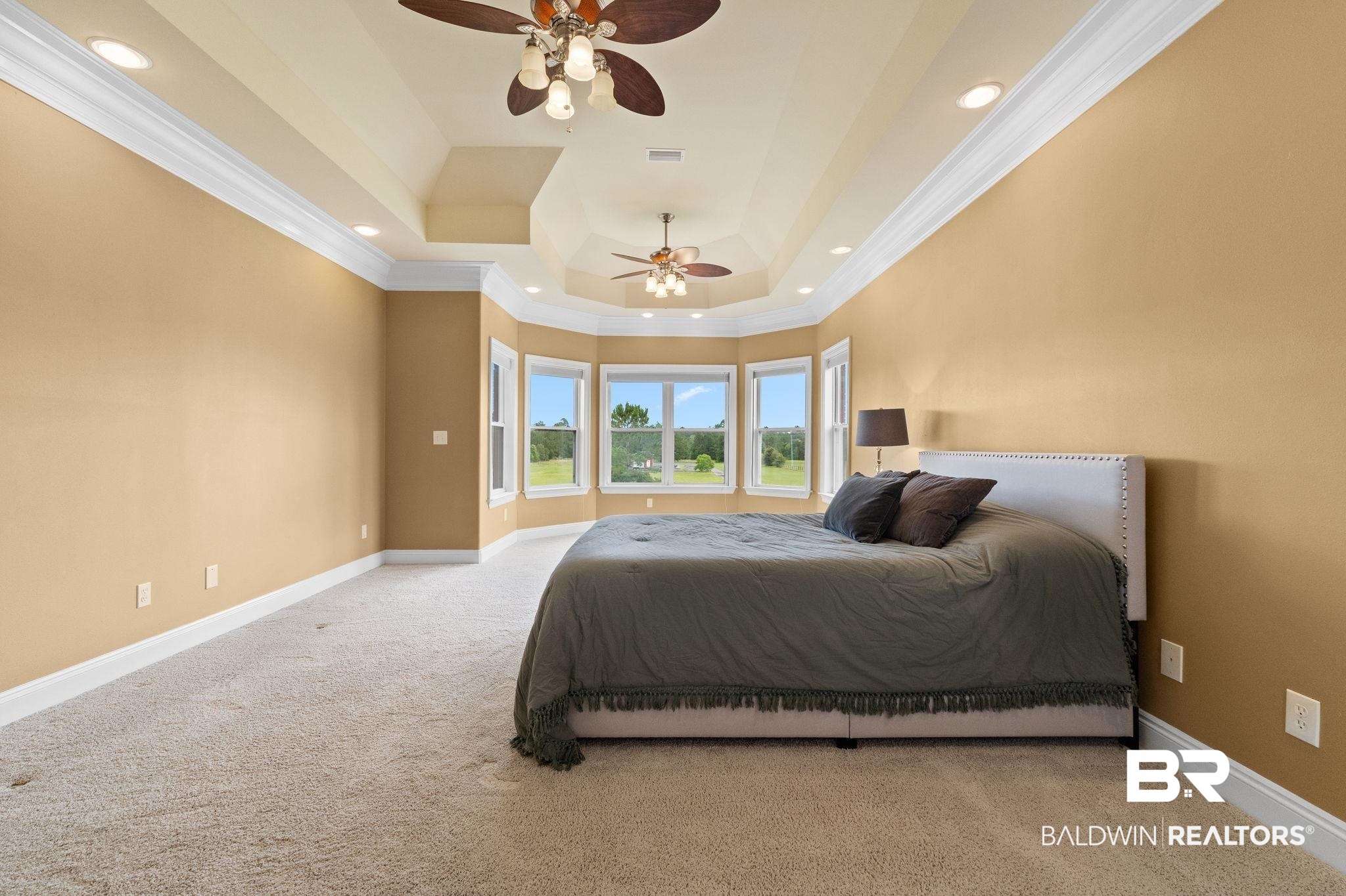 Staged bedroom with a gray bed beneath a tray ceiling and windows overlooking a green lawn.