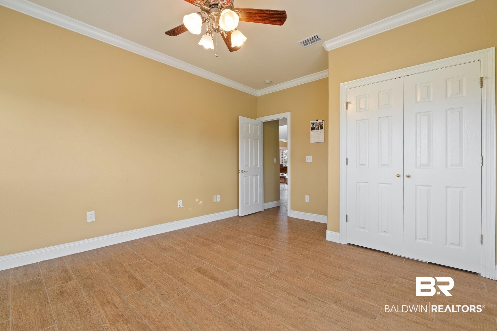 Empty bedroom with tan walls, wood-look tile floors, white closet doors, and a ceiling fan.
