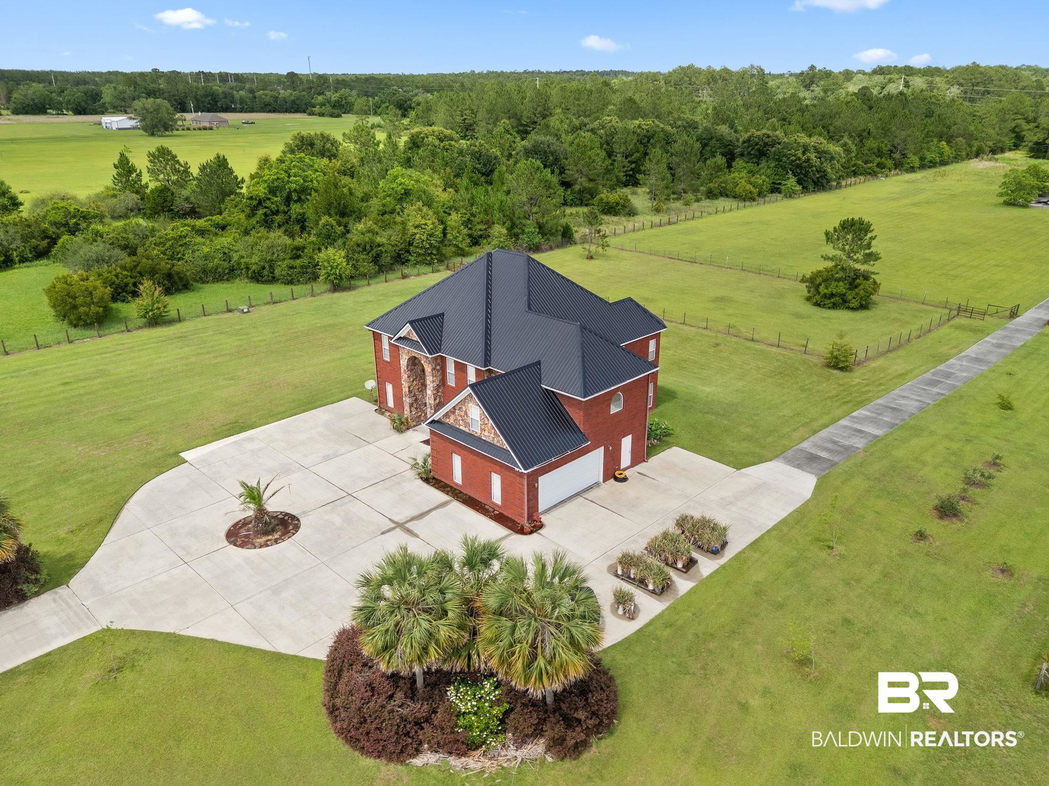 Aerial view of a red brick house with a dark metal roof, broad driveway, and expansive green lawns.