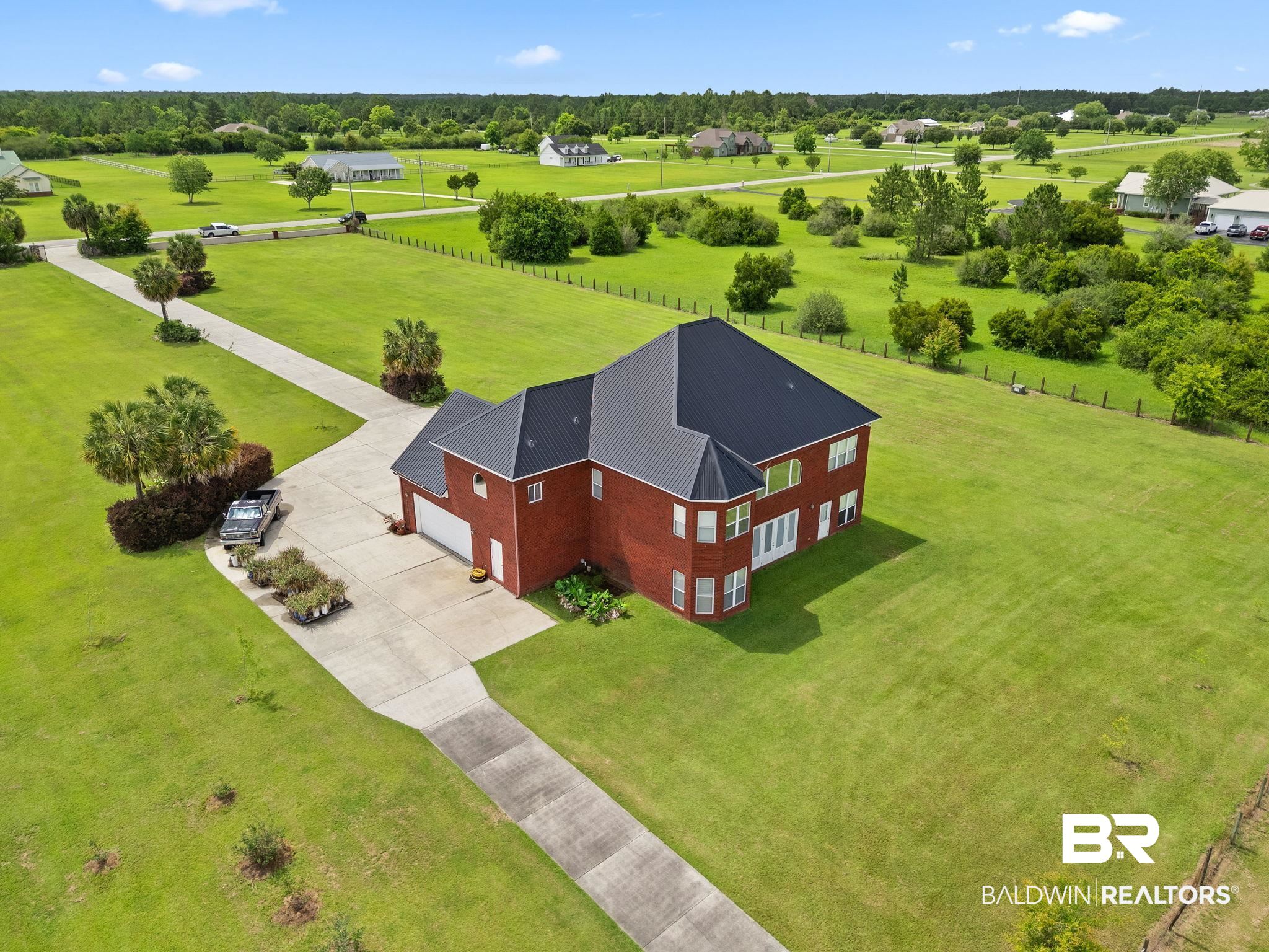 Aerial view of a red-brick home with dark metal roof, long driveway, and expansive lawn.