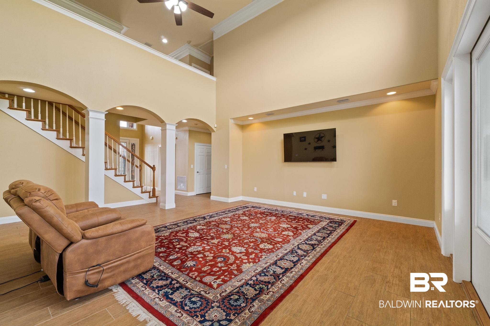 Two-story living room with curved staircase, tan recliner, and large red patterned rug.