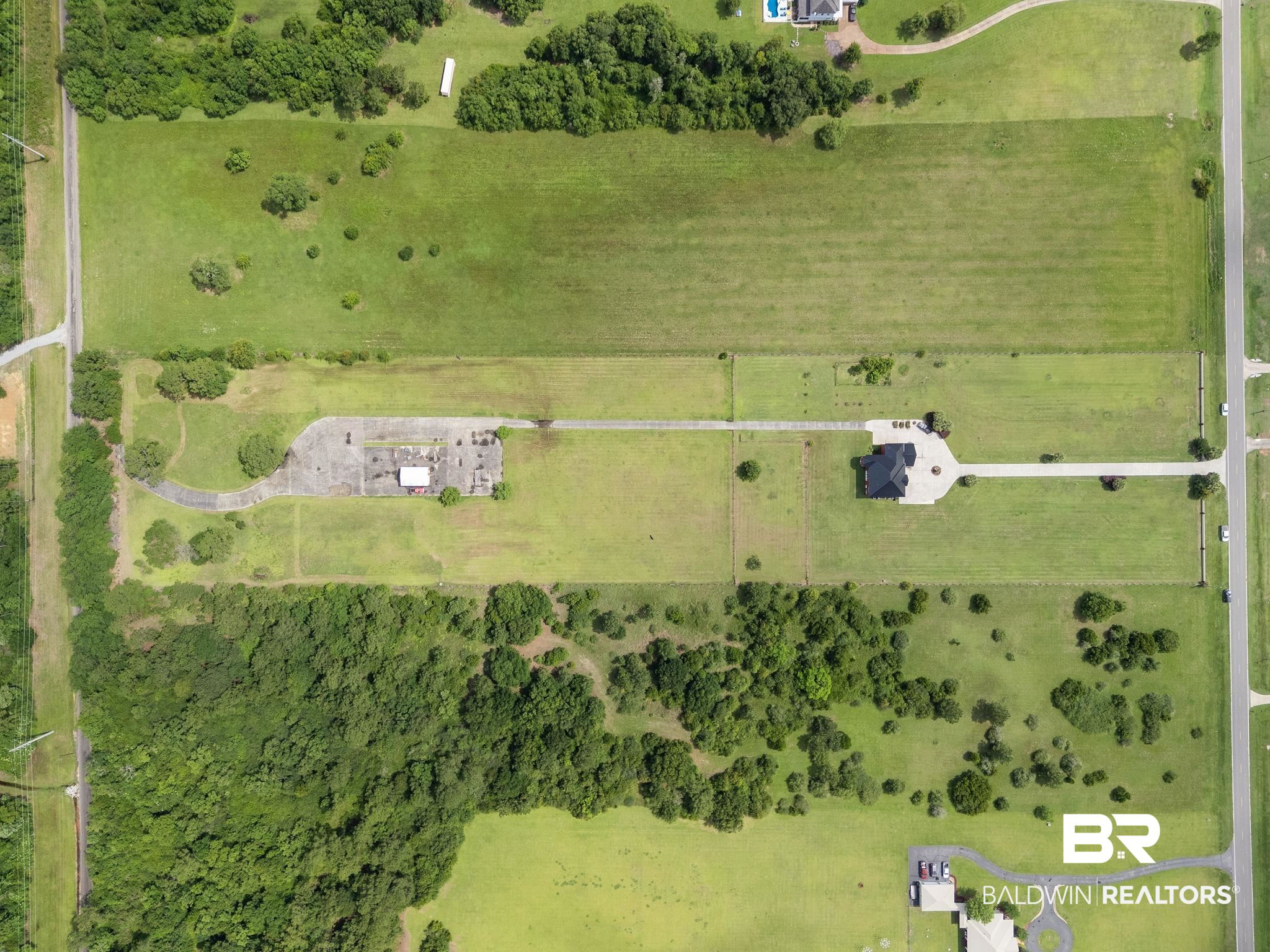 Aerial view of a spacious grassy property with a house, long driveway, and wooded areas.