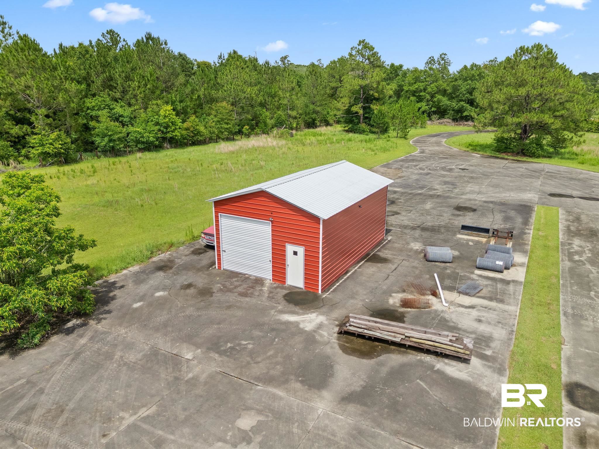 Red metal outbuilding with a white roof on a paved lot bordered by grass and trees.