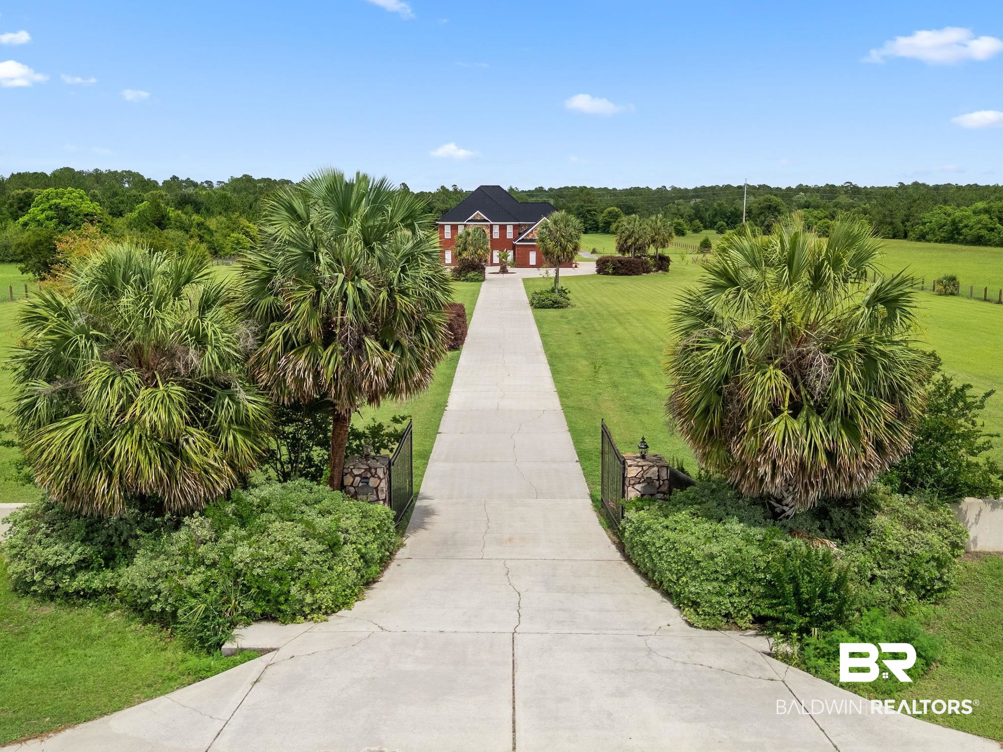 Aerial view of a red-brick house at the end of a long driveway, framed by palm trees and expansive lawns.