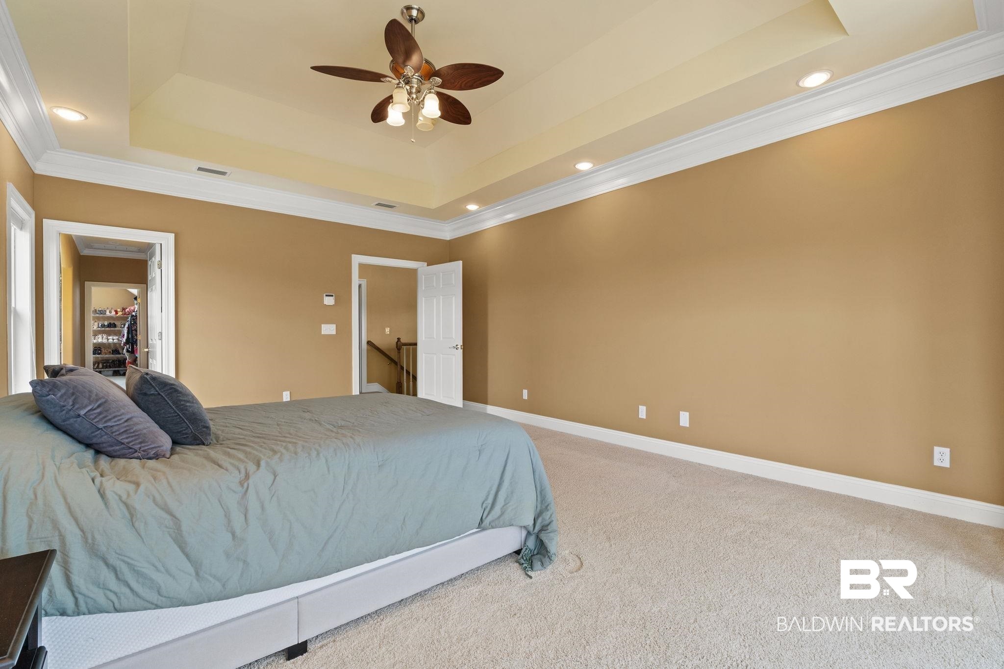 Bedroom with a gray-covered bed, tray ceiling, ceiling fan, and open doorways.