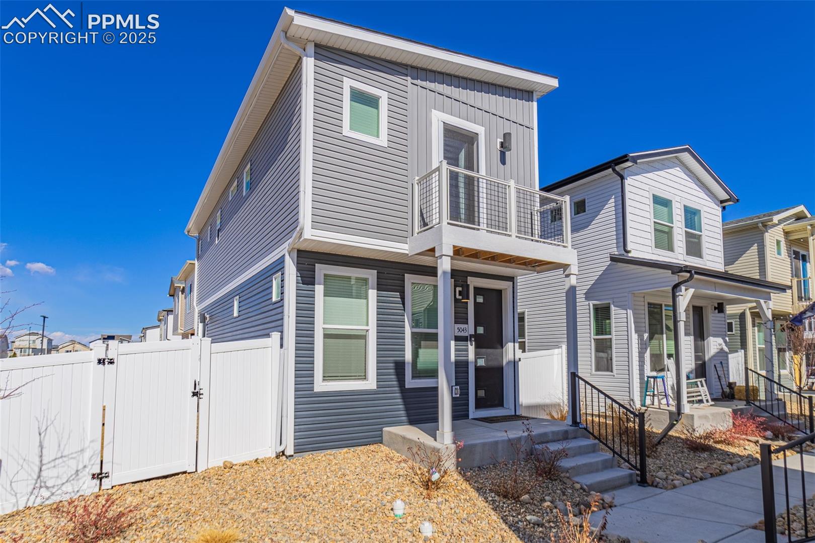 Two-story gray home with an upper balcony, white privacy fence, and rock landscaping.