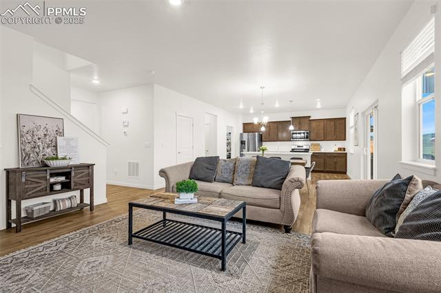 Open living room with beige sofas and patterned rug, opening into a kitchen with dark wood cabinets.