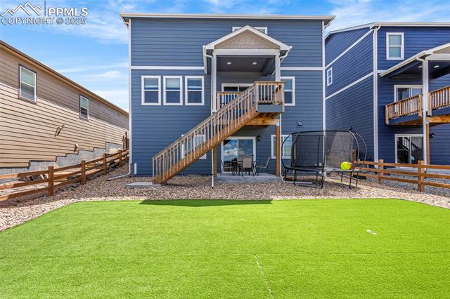 Blue two-story house with a raised deck, long staircase, artificial-turf yard, and trampoline.