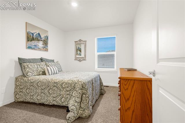 Furnished bedroom with patterned gray bedding, blue window blinds, beige carpet, and a wooden dresser.