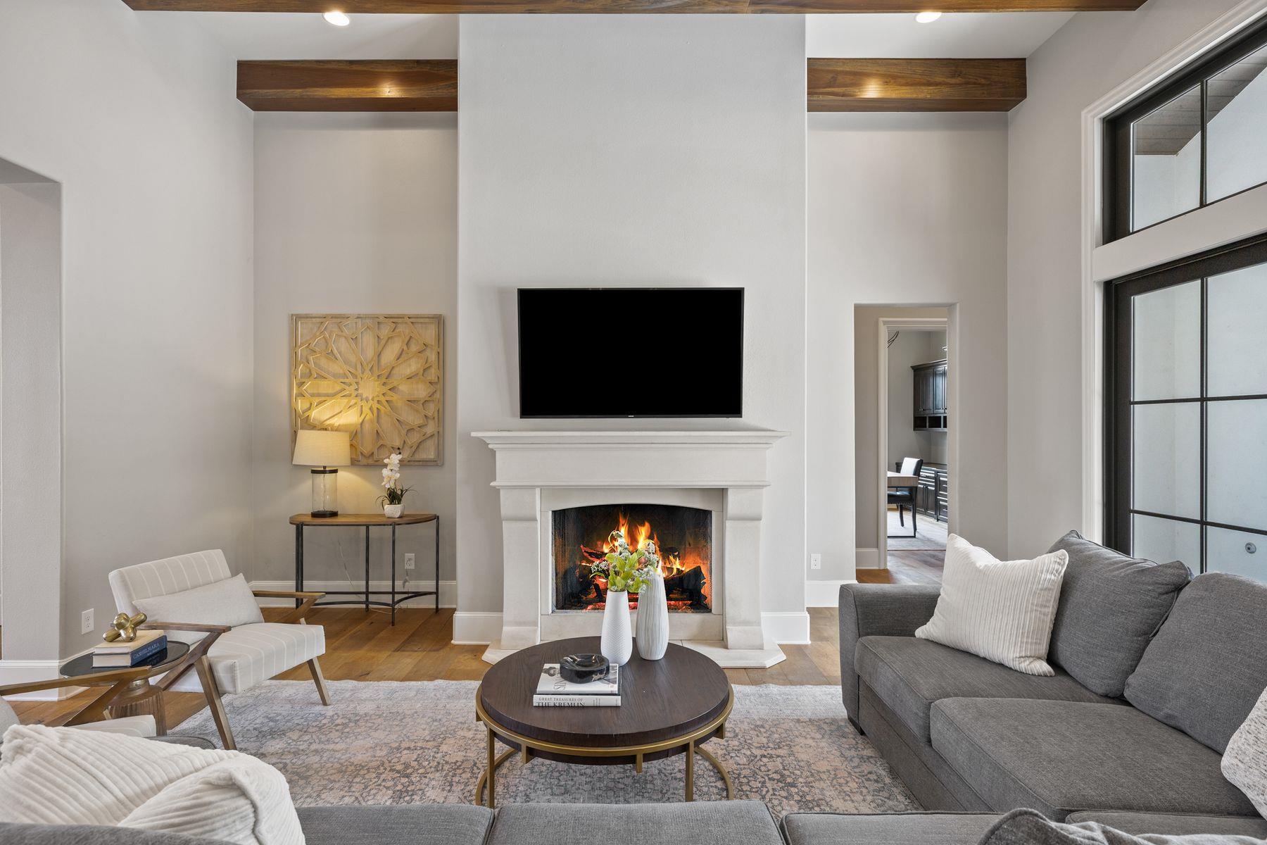 Living room with a lit fireplace, wall-mounted television, gray sofas, and tall black-framed glass doors.