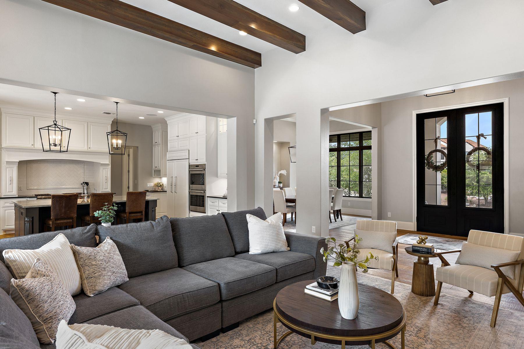 Open-plan living room with exposed wood beams, gray sectional, white kitchen, and black French doors.