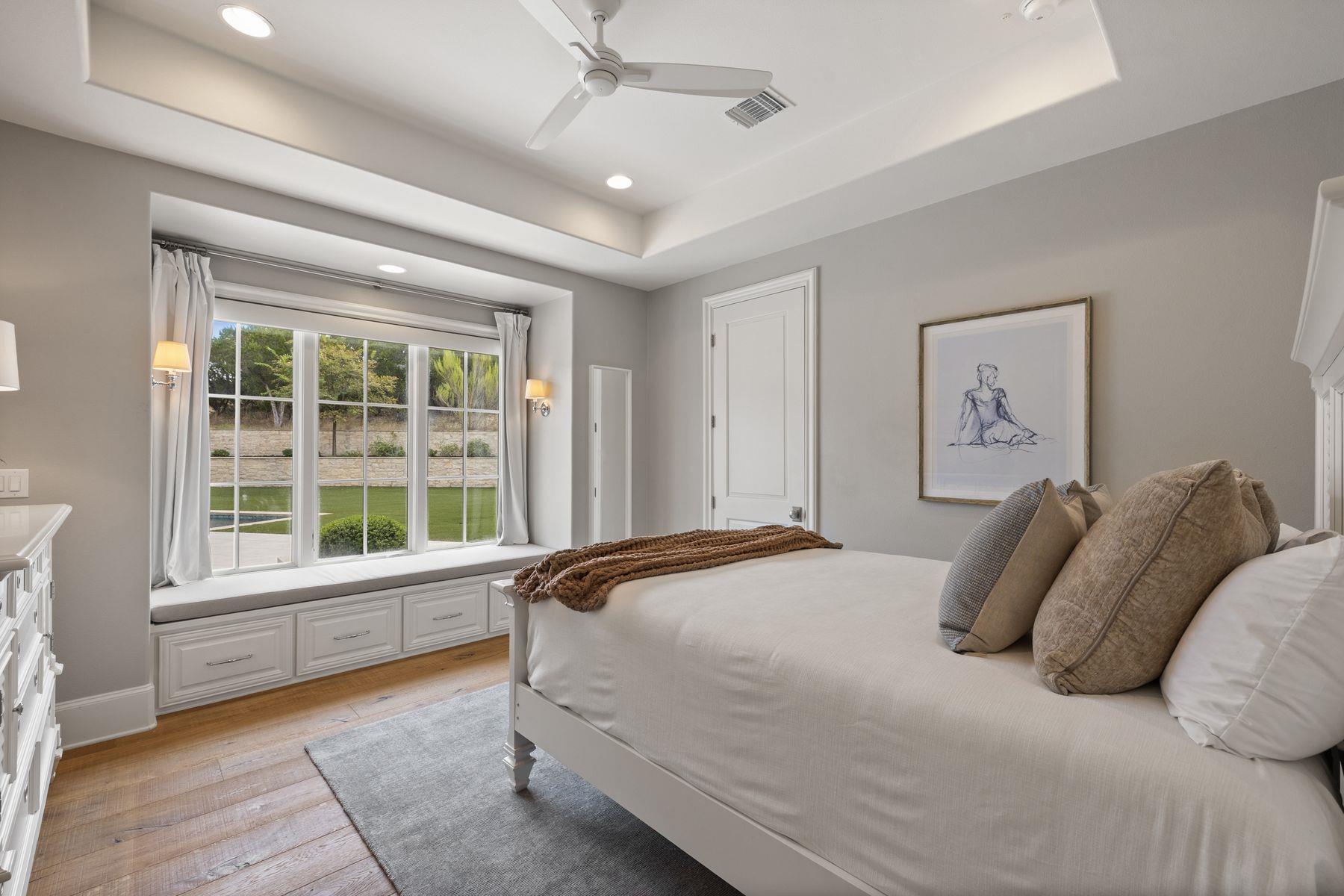Bedroom with a white bed and window-seat drawers overlooking a lawn and glimpse of a pool.