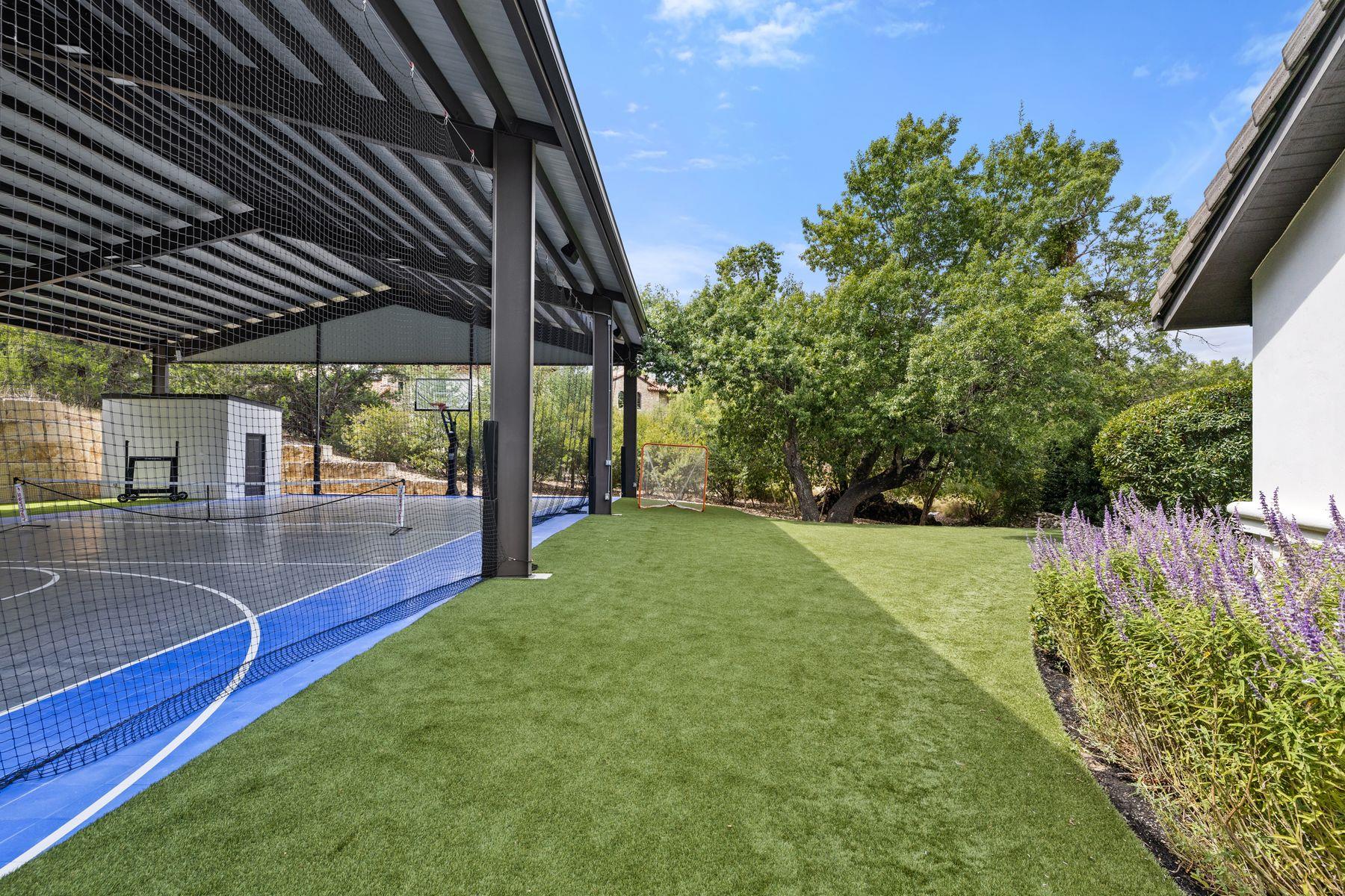 Covered basketball court beside an artificial-turf lawn, soccer goal, mature trees, and purple flowers.