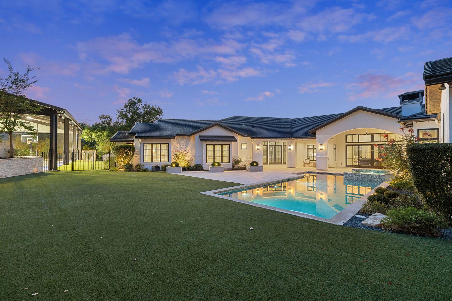 Home’s rear yard at dusk, with an illuminated pool, broad lawn, covered patio, and enclosed basketball court.