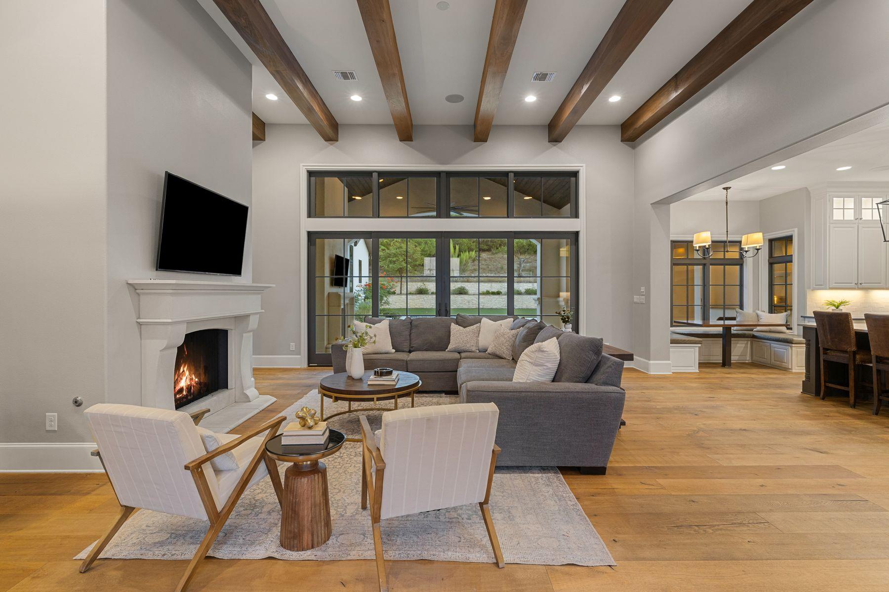 Living room with exposed wood beams, gray sectional, fireplace, and large windows overlooking the yard.