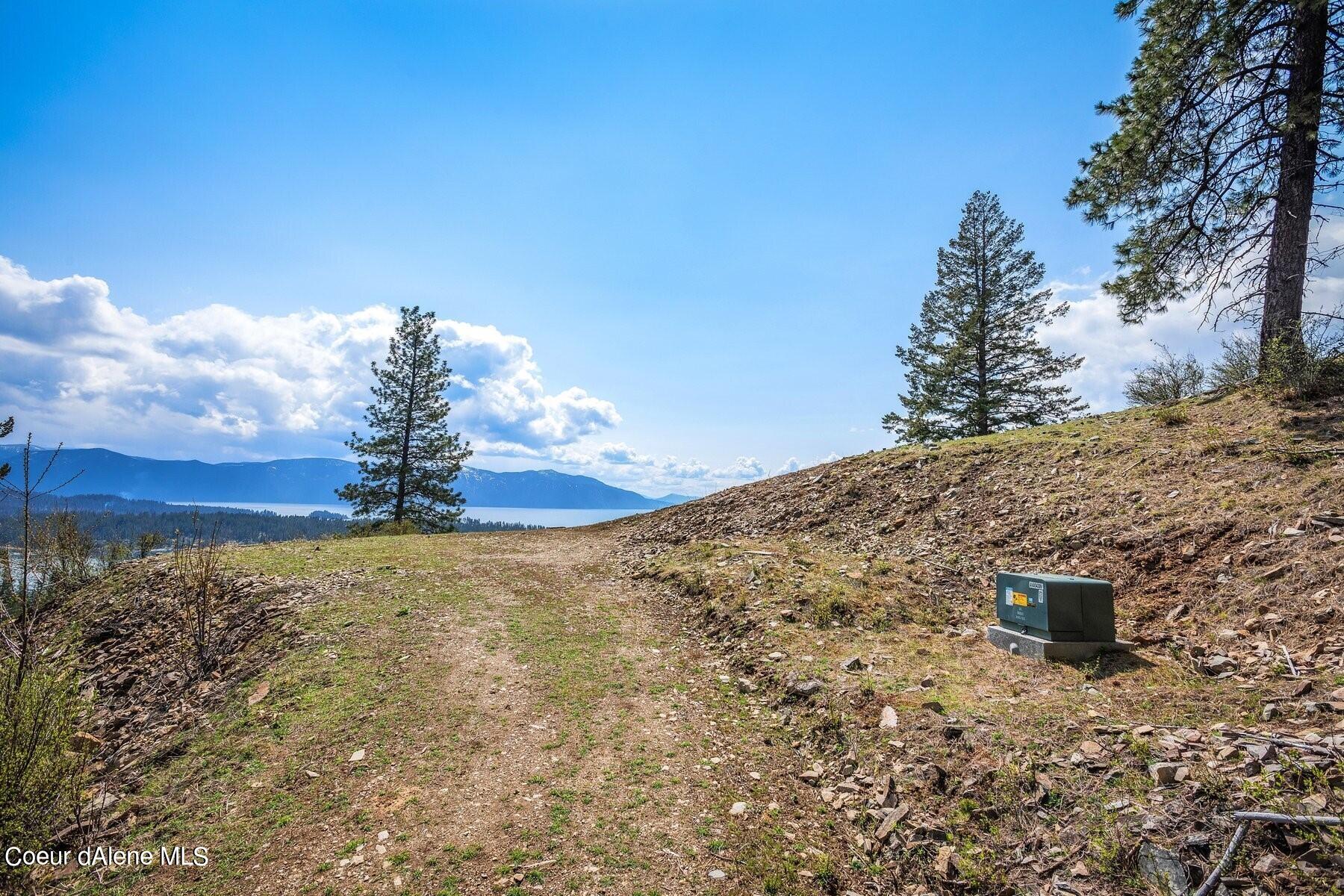 Vacant hillside lot overlooking a lake and distant mountains, with pine trees and a rocky dirt track.