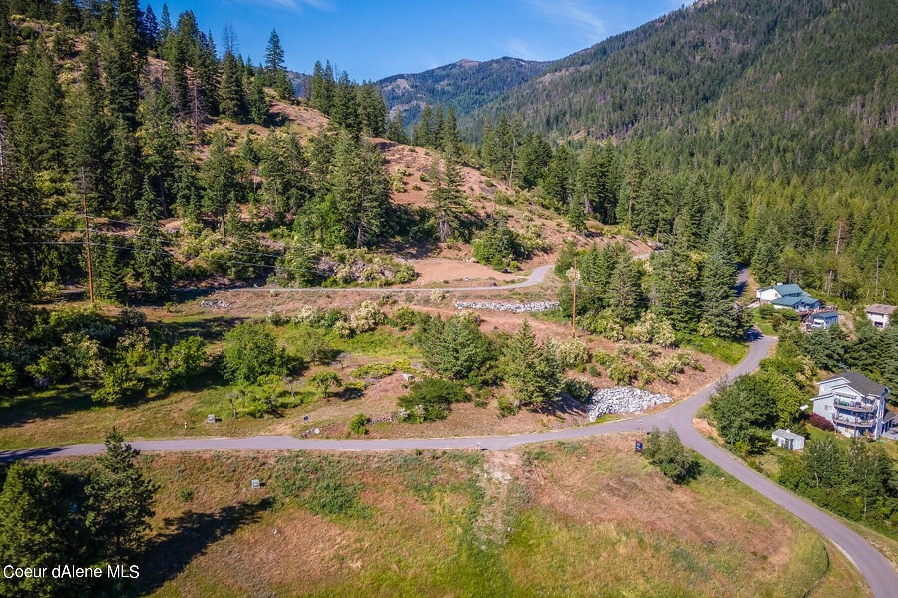 Aerial view of a cleared hillside lot beside a curving road, surrounded by forested mountains.
