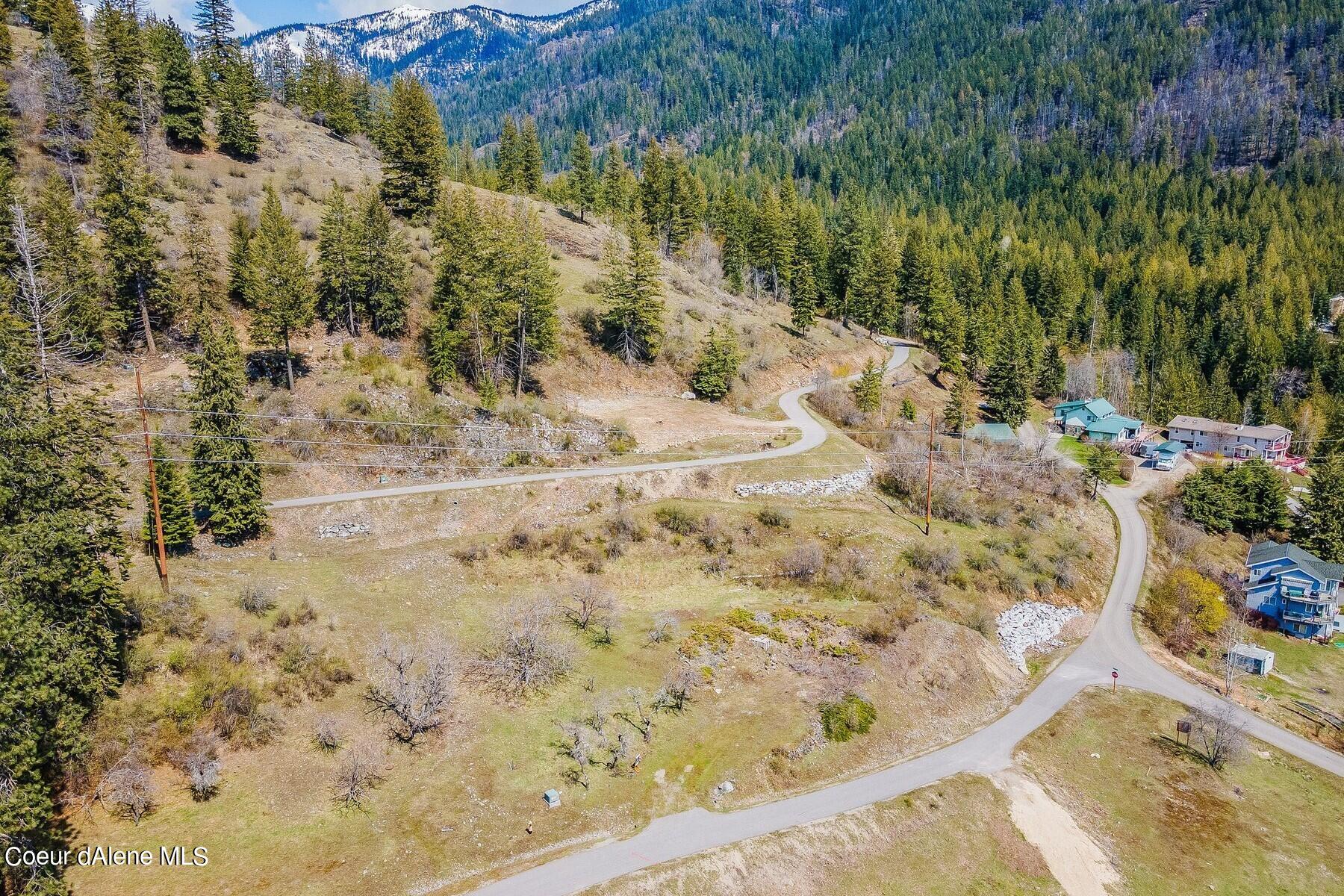 Vacant hillside lot beside a winding road, with forested mountains and nearby homes.