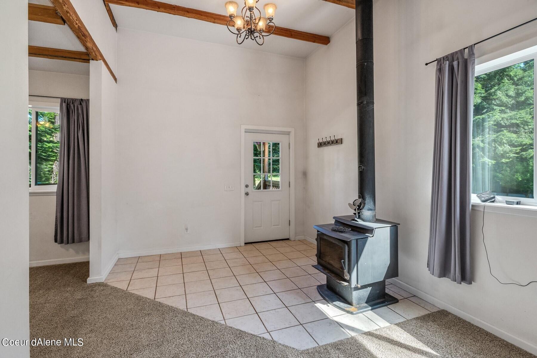 Unfurnished room with a freestanding wood stove, exposed ceiling beams, and windows overlooking trees.