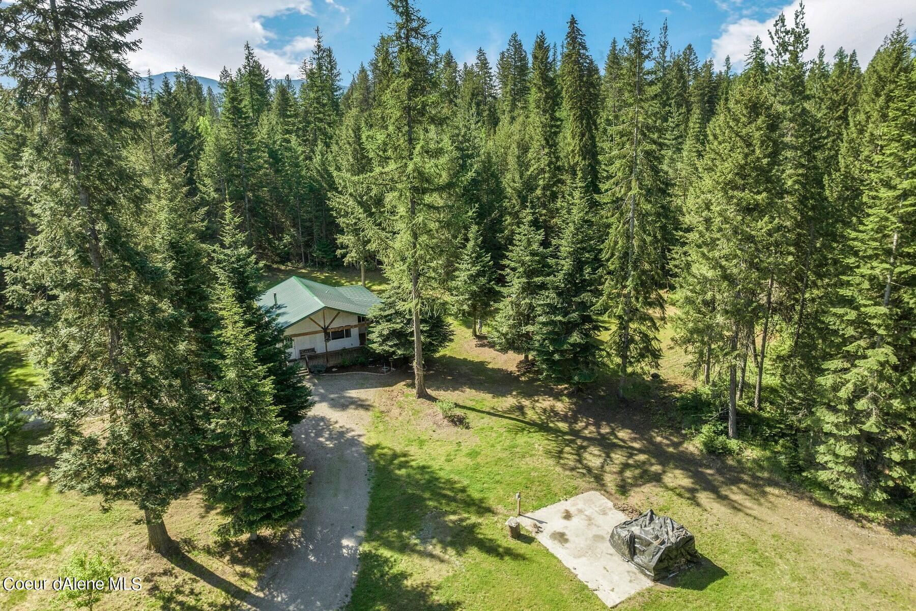 Aerial view of a green-roofed cabin in a forest clearing with a gravel driveway.