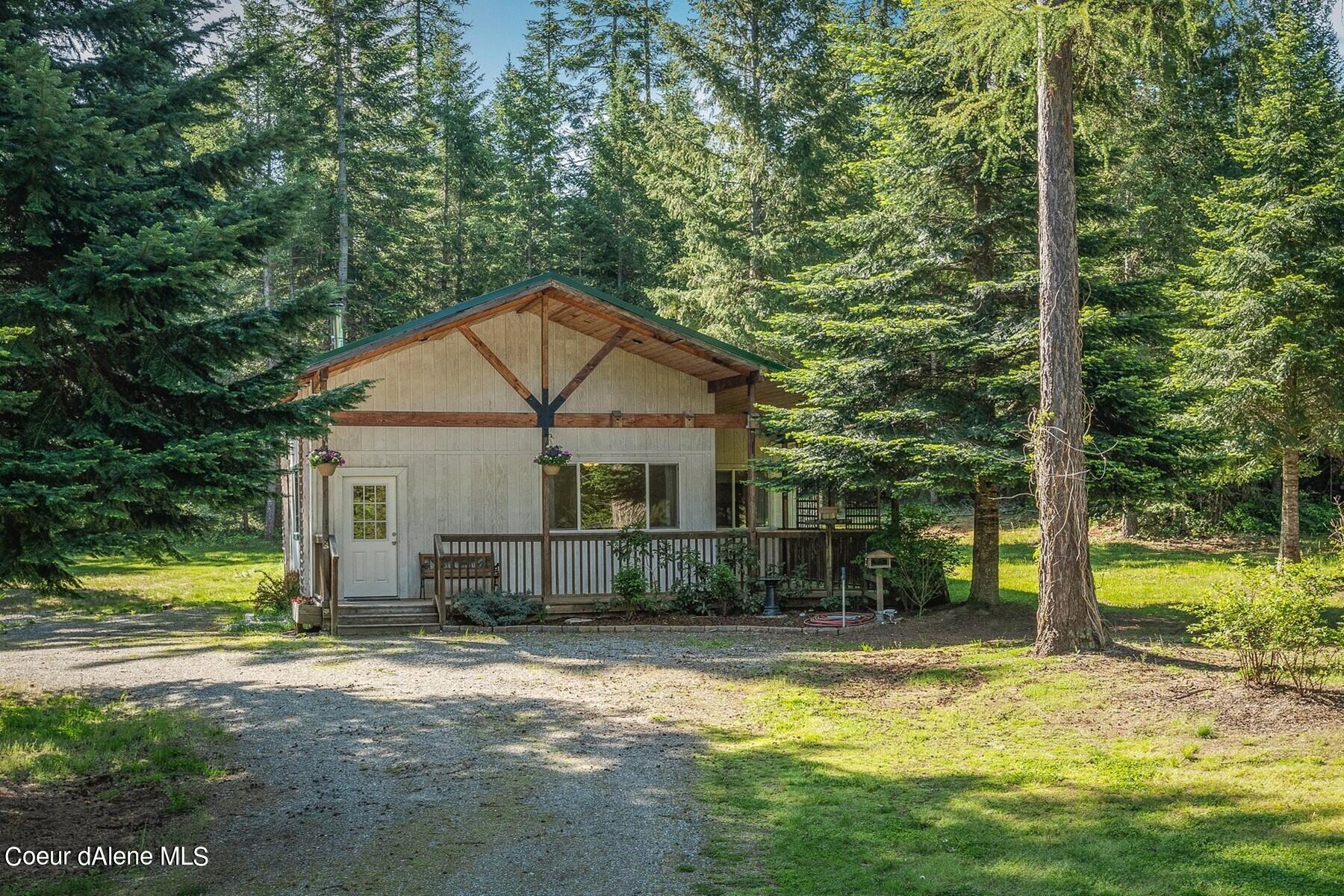 Cottage with a covered porch nestled among tall trees, with a gravel driveway in front.