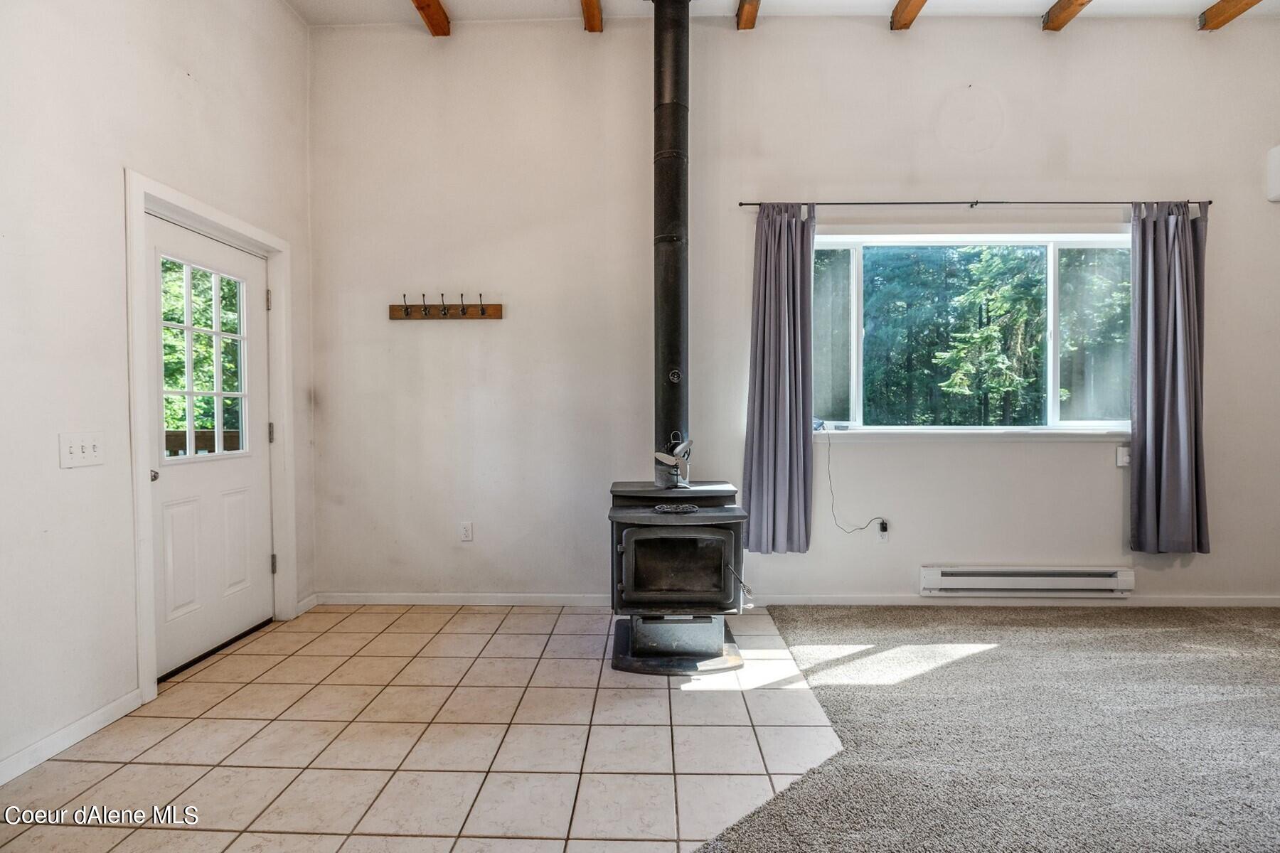 Unfurnished living room with a freestanding wood stove, exposed beams, and a large window overlooking trees.