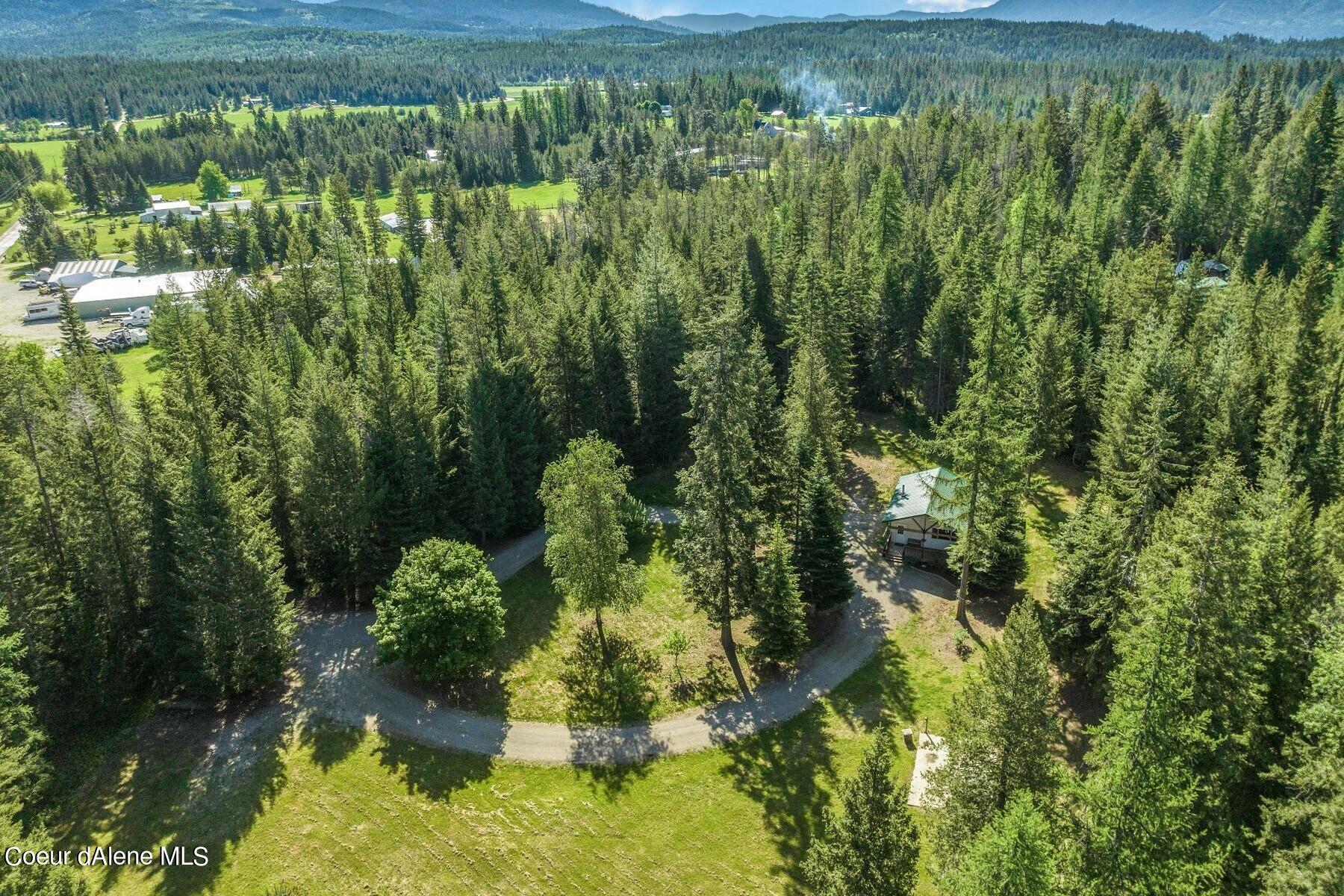 Aerial view of a wooded property with a small building and gravel drive, surrounded by evergreen forest and distant mountains.