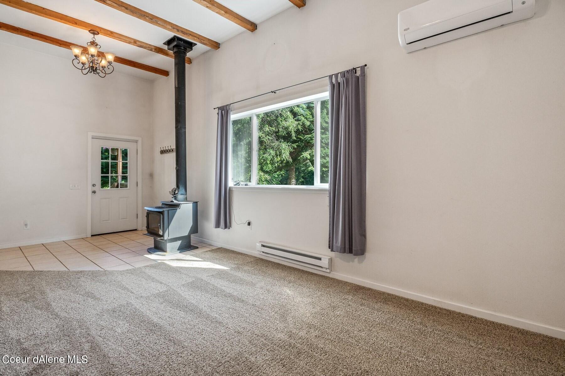 Unfurnished living room with a freestanding wood stove, exposed beams, and a large window overlooking trees.