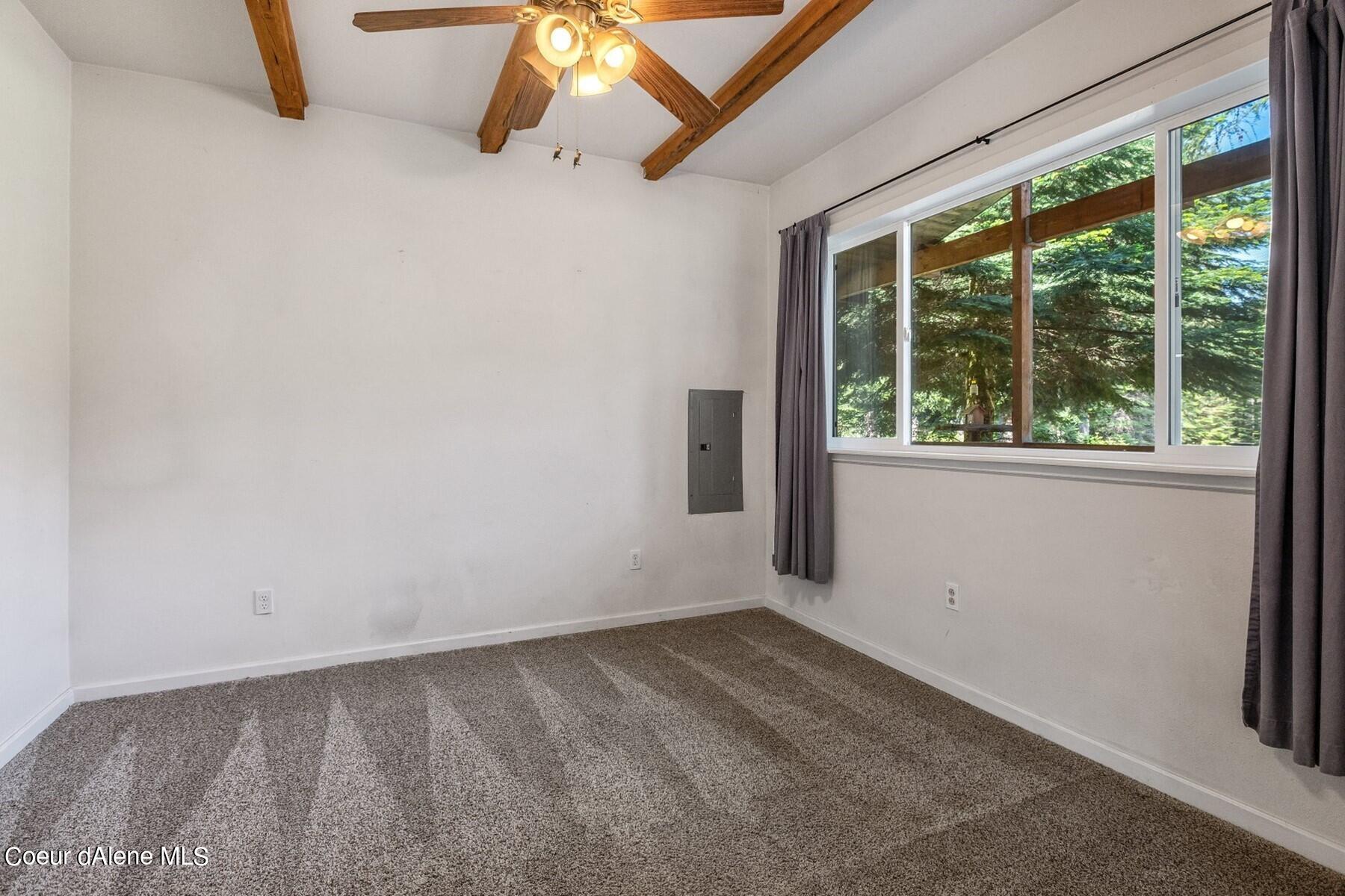 Empty carpeted room with exposed ceiling beams and a wide window overlooking trees.
