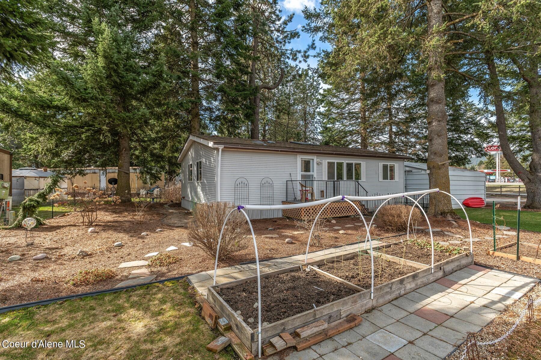 Single-story home with a rear deck, paved patio, and raised garden beds beneath tall trees.