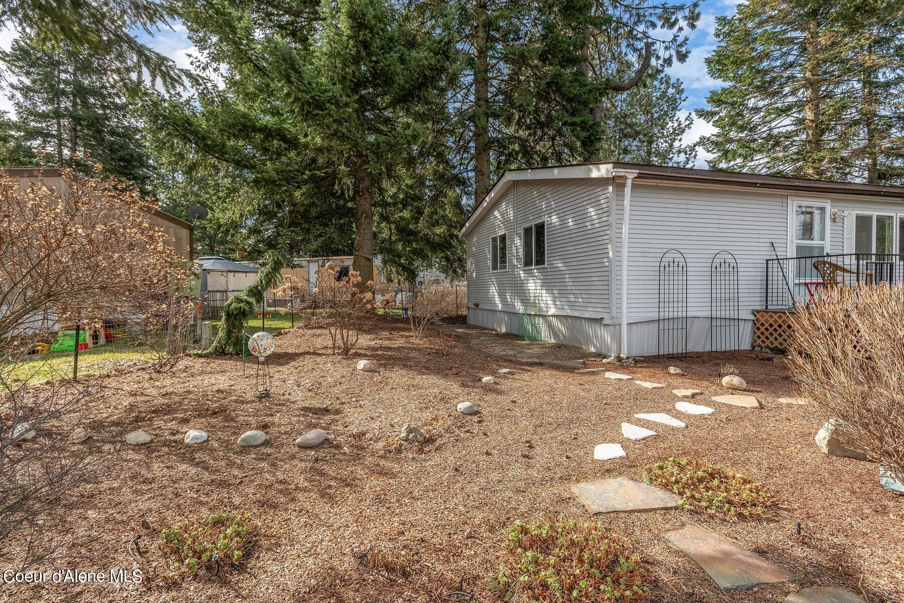 Gray manufactured home beside a mulched yard with stepping stones and mature evergreens.