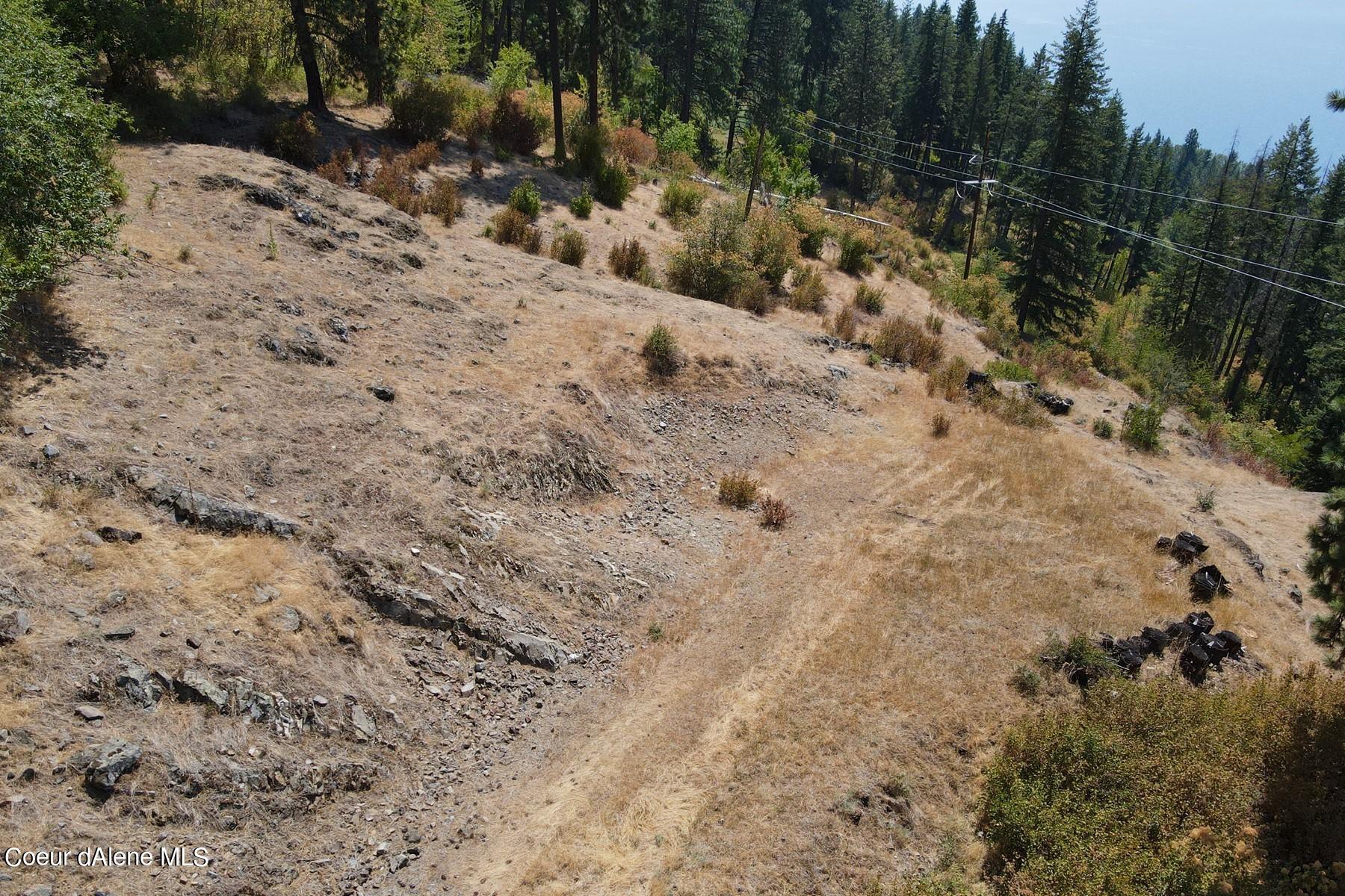 Aerial view of a dry, rocky hillside lot bordered by forest, with power lines and water in the distance.