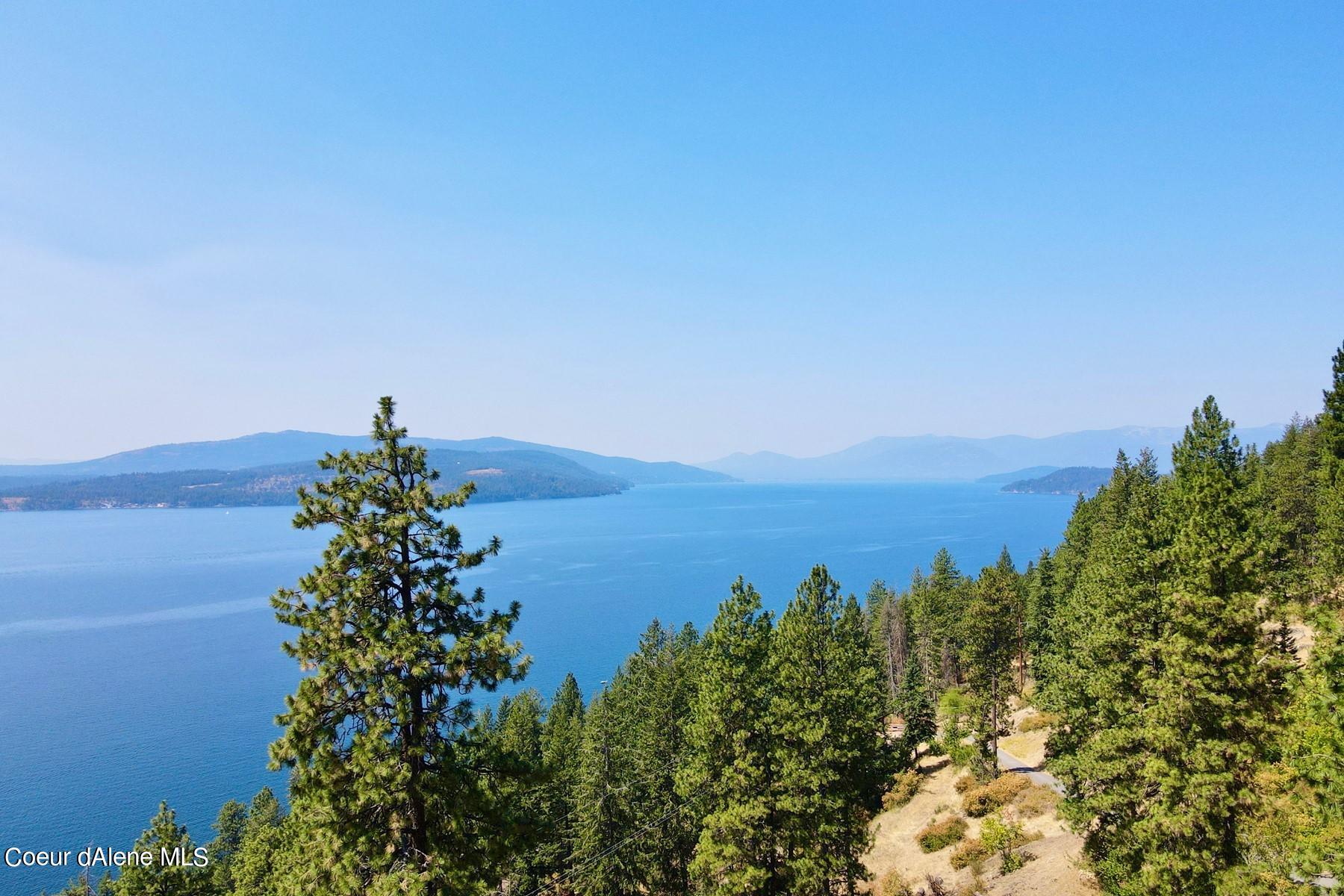 Blue lake and distant mountains viewed over pine-covered hillsides, with a road winding through the foreground.