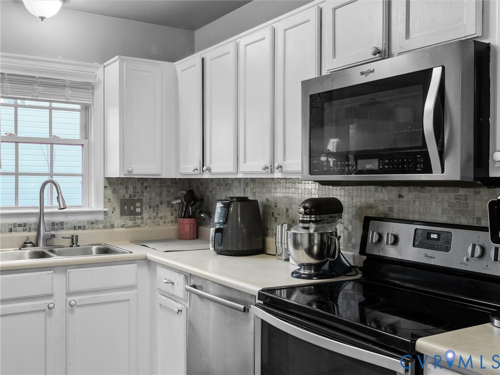 Kitchen with white cabinets, gray mosaic backsplash, window-side sink, and stainless steel appliances.