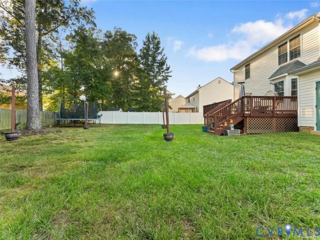 Backyard with a wide lawn, mature trees, white privacy fence, trampoline, and raised wooden deck.