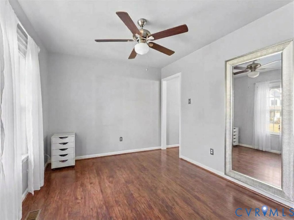 Mostly empty bedroom with reddish wood floors, sheer curtains, a ceiling fan, and a large mirror.
