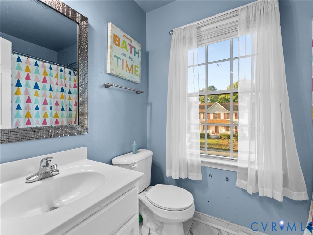 Blue bathroom with a white vanity, toilet, mosaic-framed mirror, and sheer-curtained window.