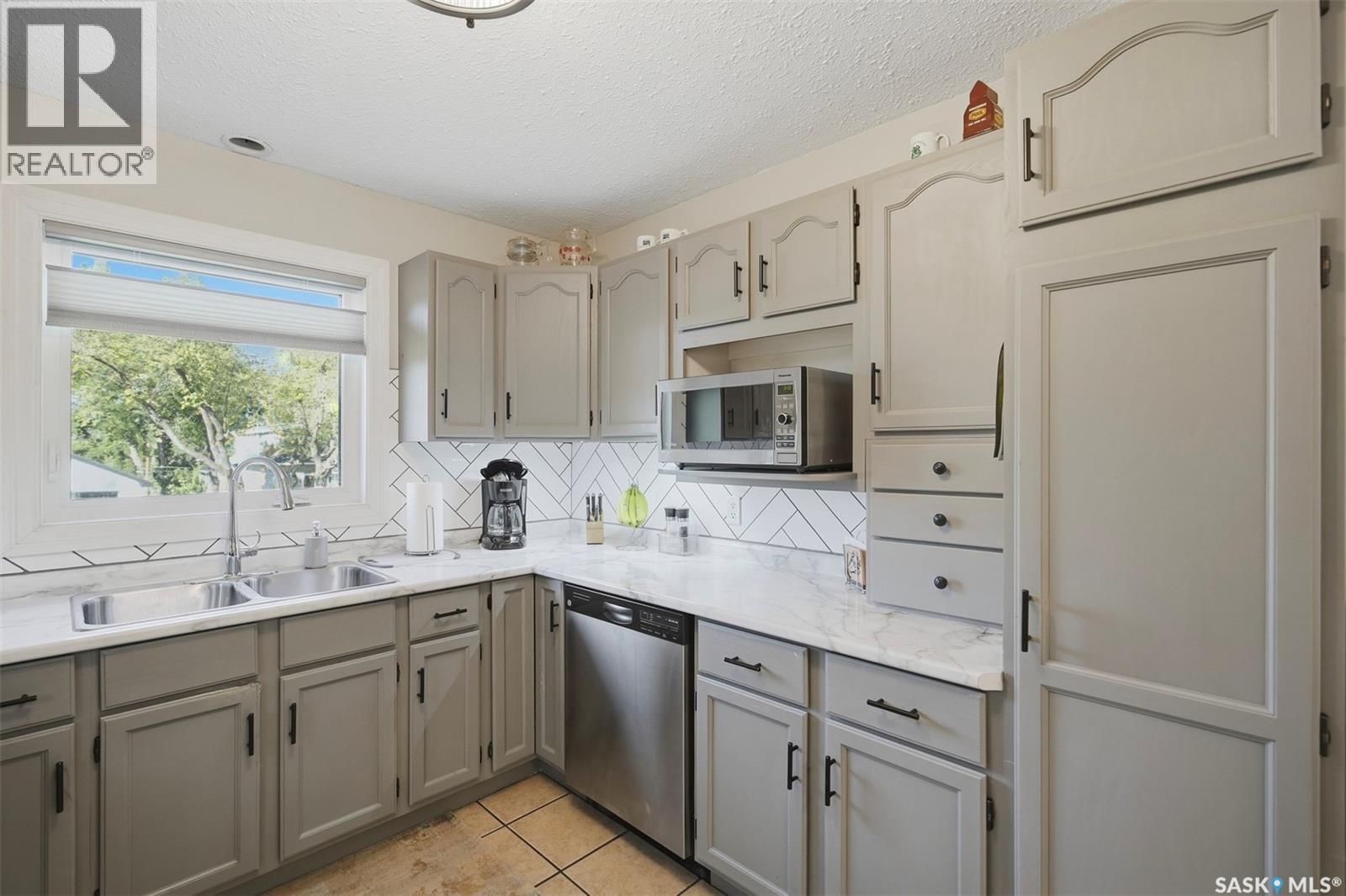 Kitchen with gray cabinets, marble-look counters, white tile backsplash, and a double-basin sink beneath a window.