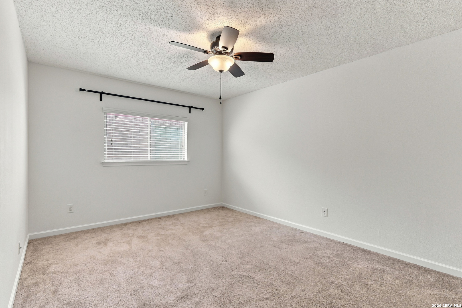 Empty bedroom with beige carpet, a window with blinds, and a ceiling fan with light.